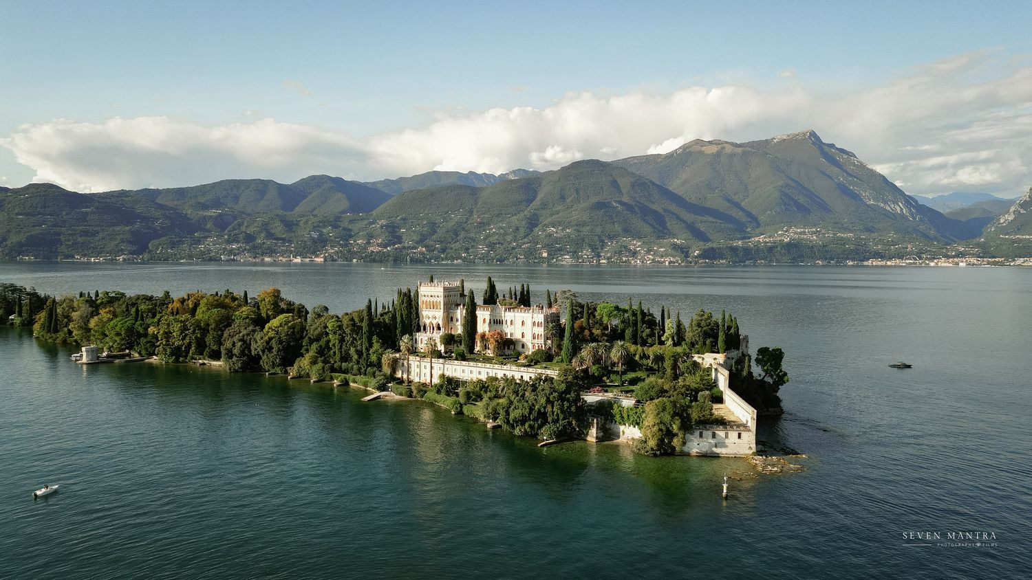Aerial view of Isola del Garda castle surrounded by Lake Garda with mountains in the background.