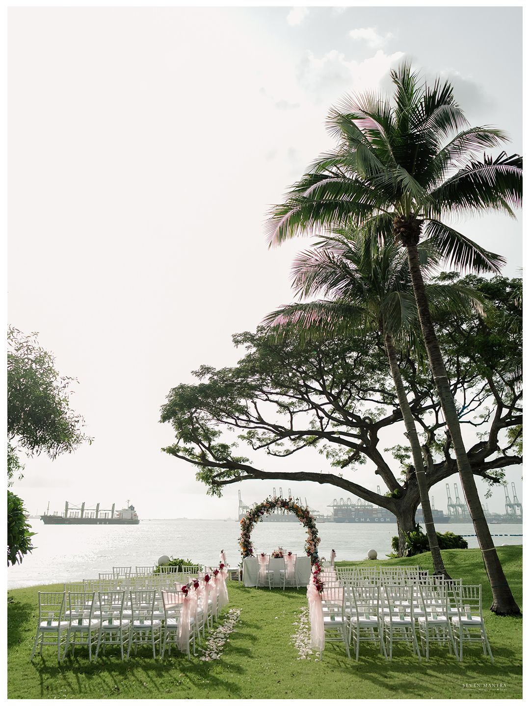 Elegant outdoor wedding ceremony setup with floral arch, white chairs, and waterfront view.