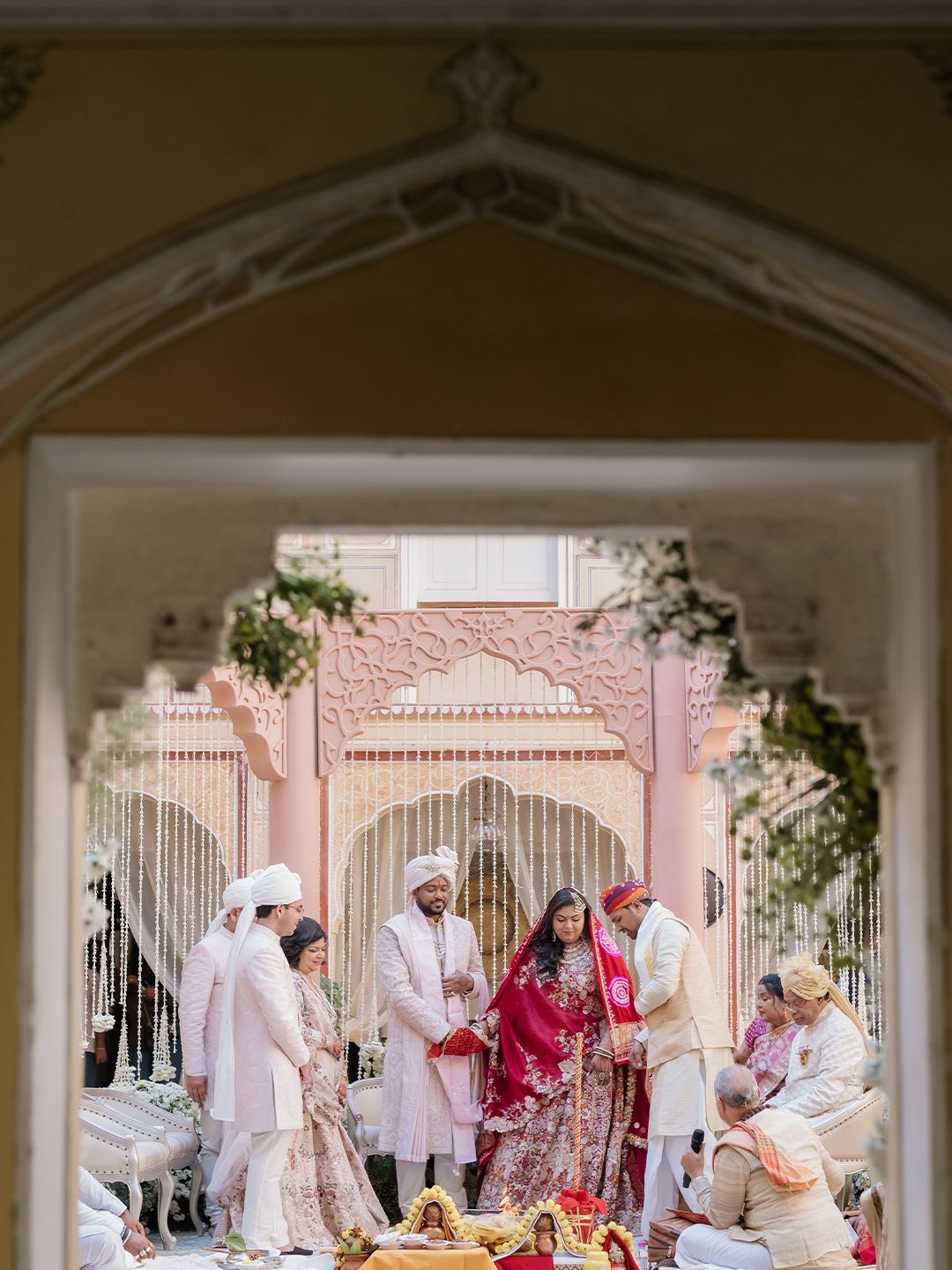 Traditional Indian wedding ceremony viewed through ornate archway at elegant pink palace venue.