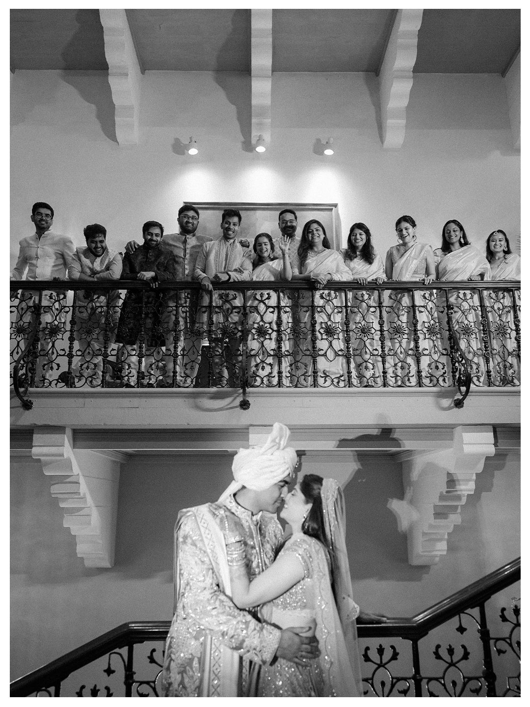 Black and white photo of Indian newlyweds kissing on stairs while guests watch from balcony above.