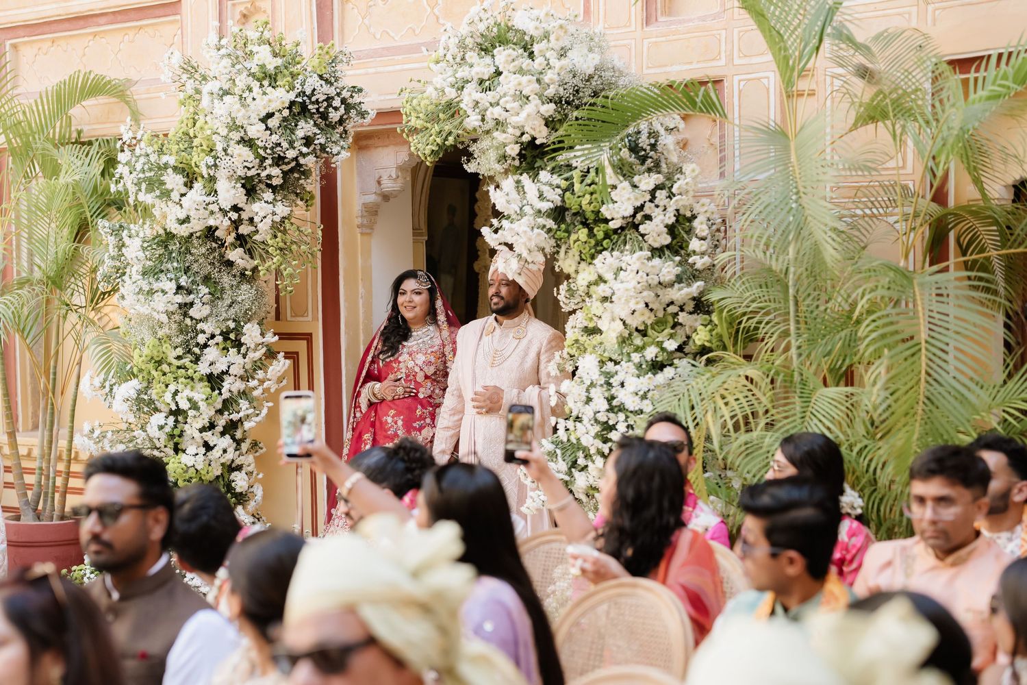 Indian bride and groom entering ceremony under lush white floral arch surrounded by wedding guests.