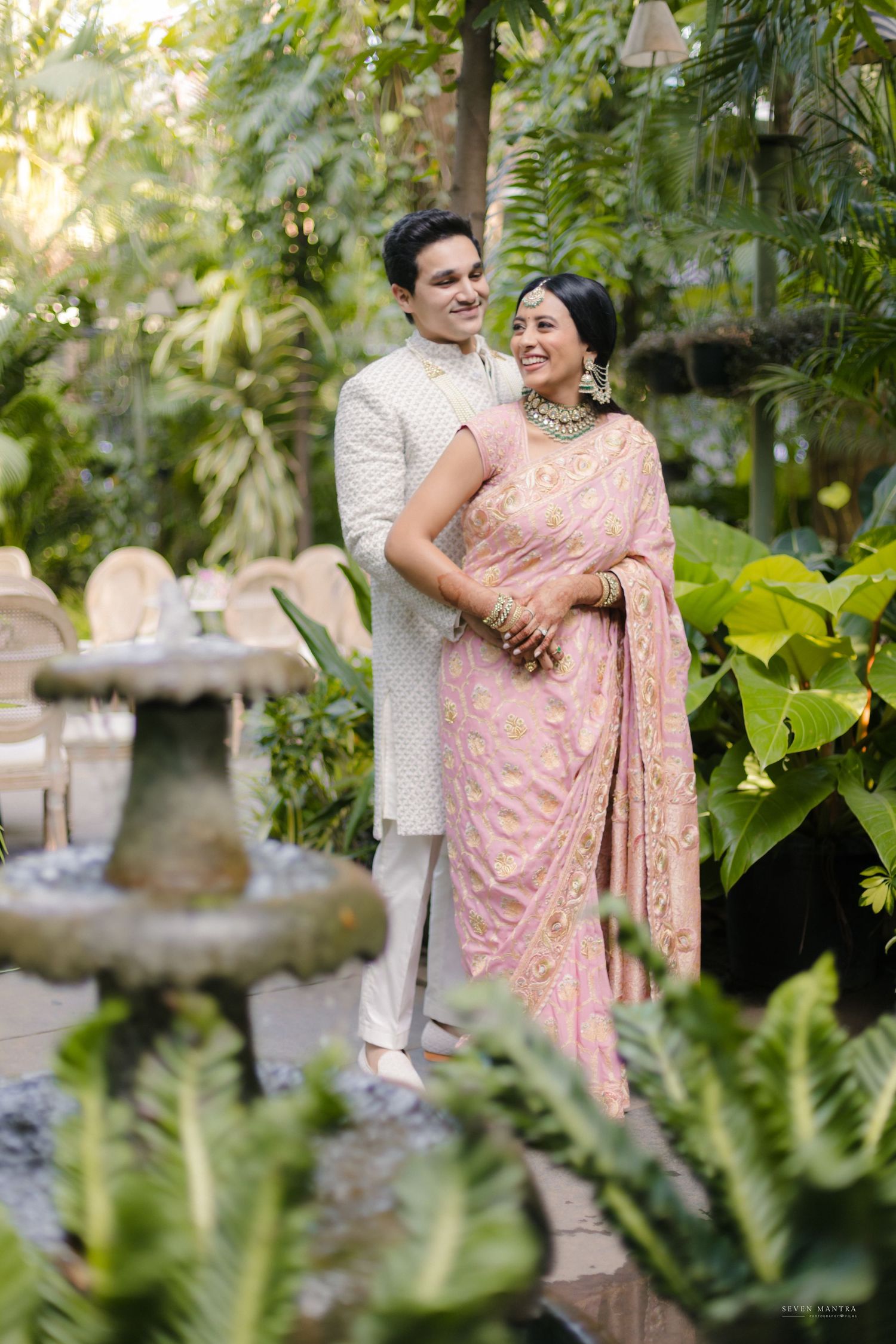 Indian couple in traditional attire pose near a garden fountain surrounded by lush tropical greenery.