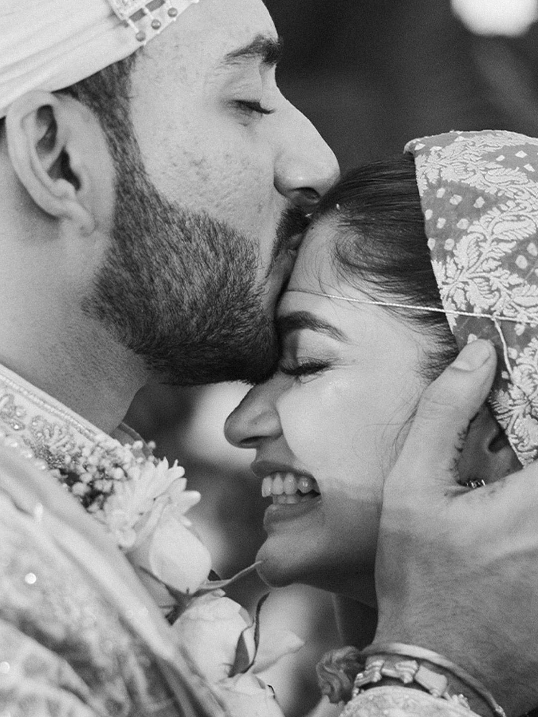 Intimate black and white close-up of groom kissing bride on forehead during Indian wedding ceremony.