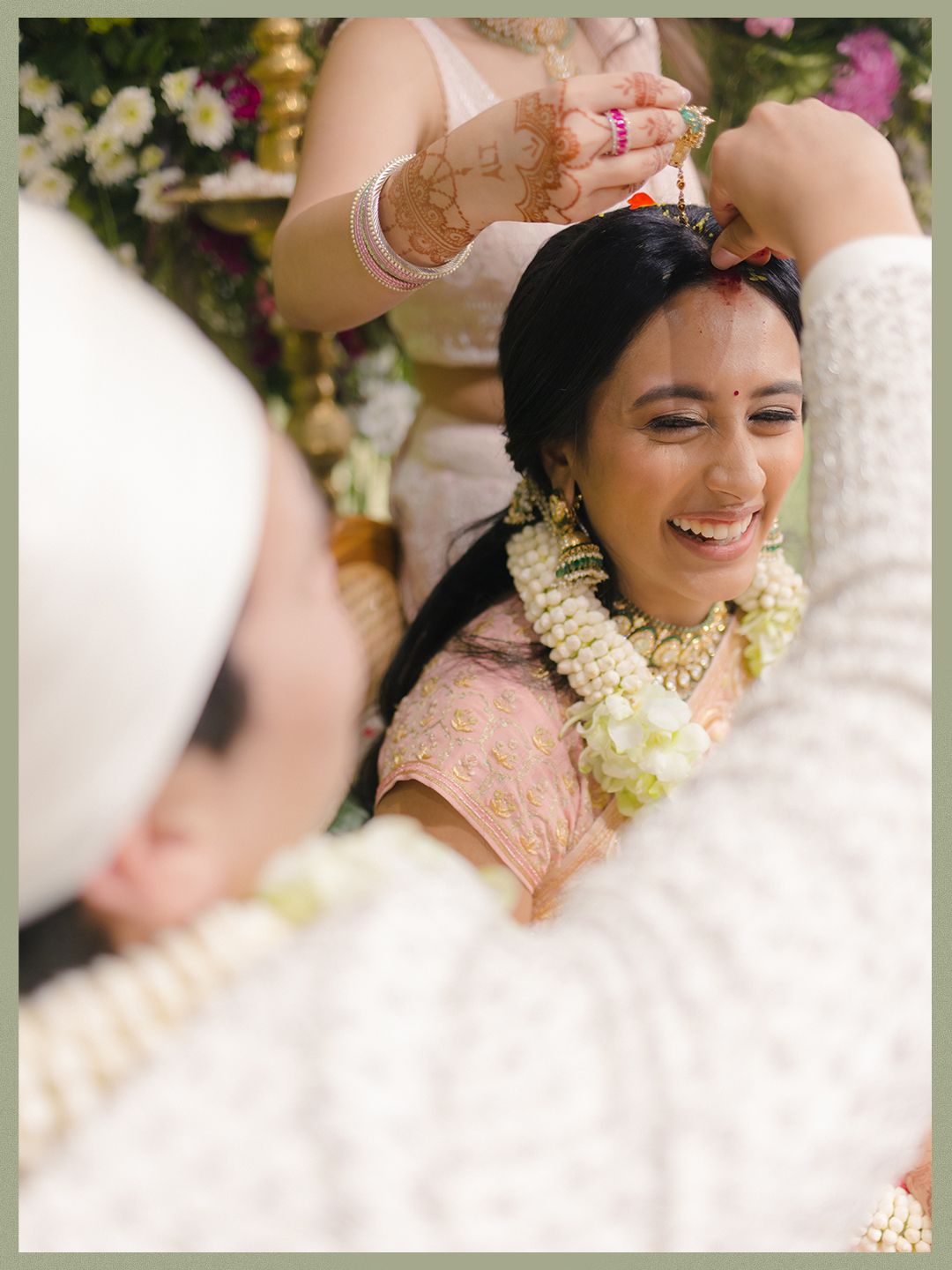 Indian bride smiling during sindoor ceremony in pink lehenga with floral garland at Hindu wedding.