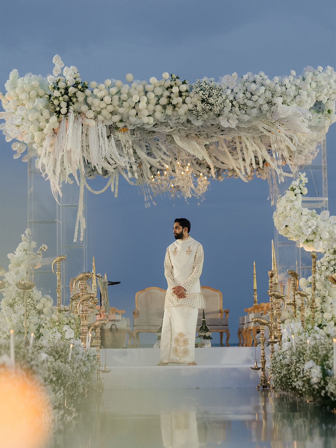 Groom in ivory sherwani stands beneath an elaborate white floral wedding mandap at twilight ceremony.