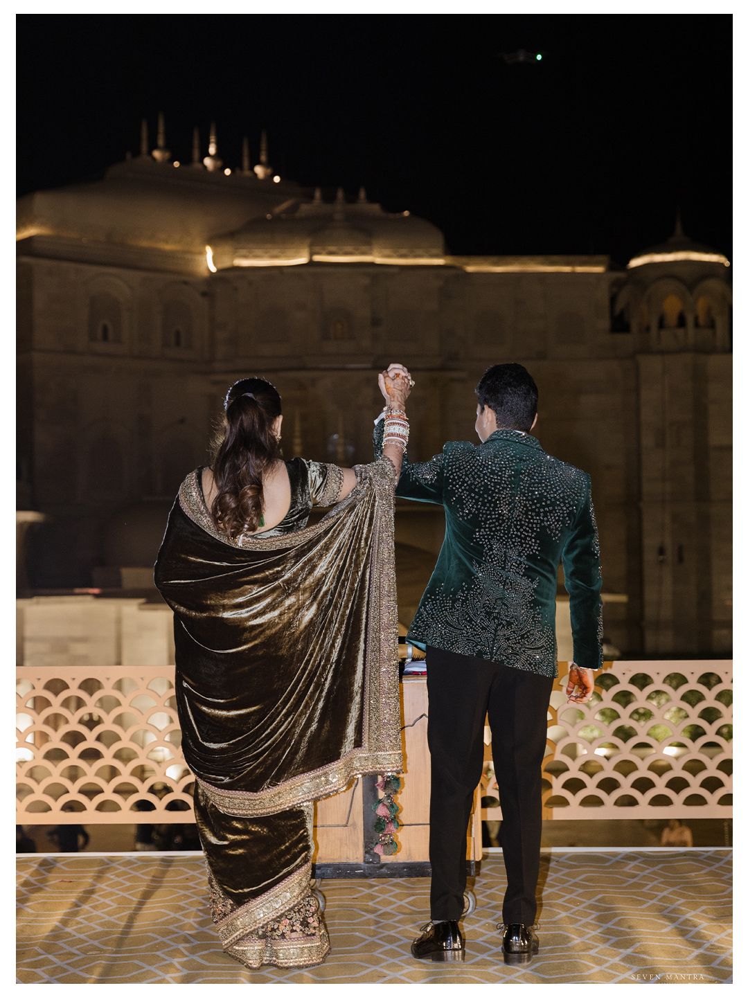 Bride in golden saree and groom in teal embroidered jacket hold hands before a lit Indian palace.