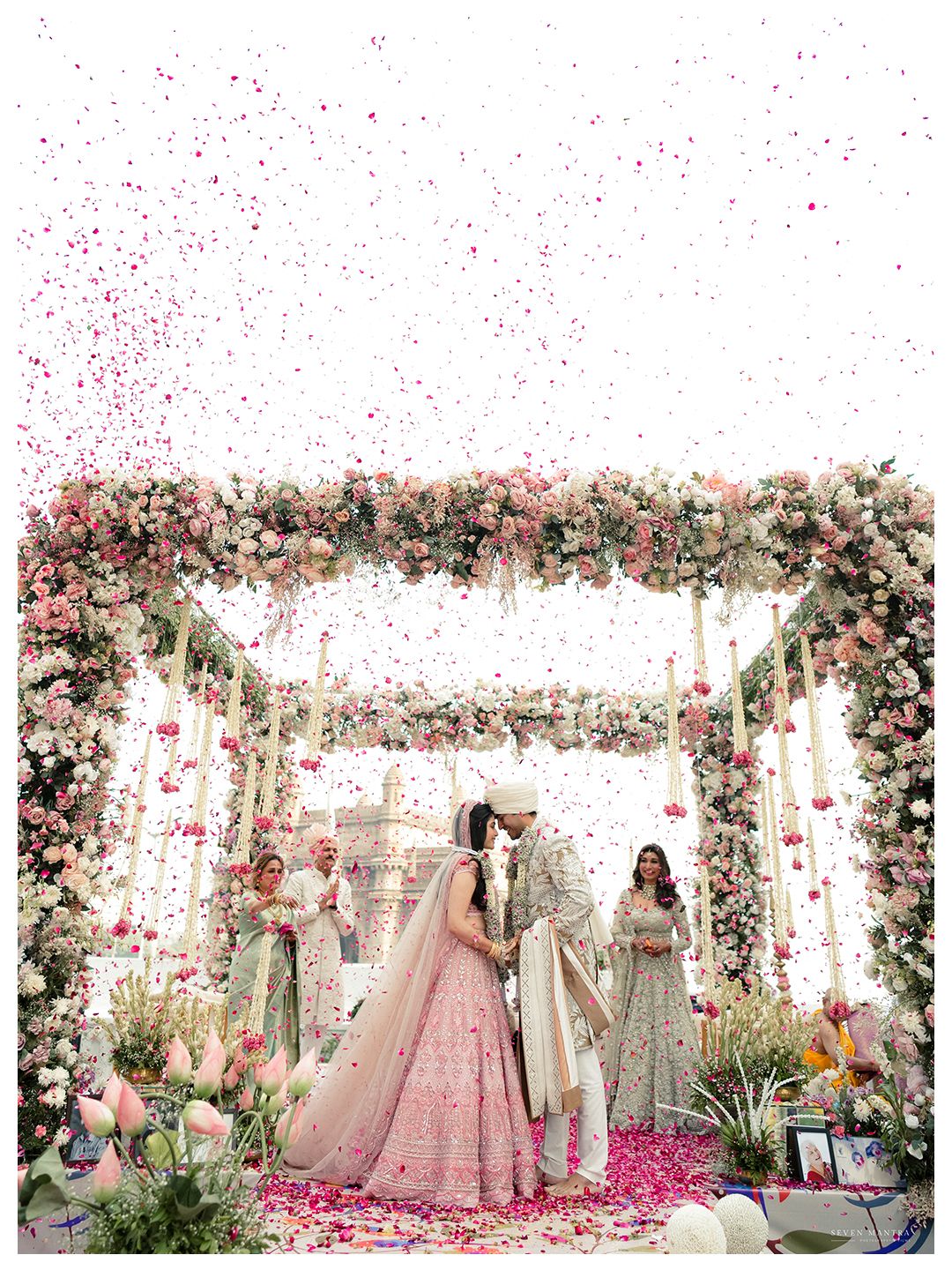 Indian wedding couple kissing under a stunning floral arch with pink rose petals raining down.