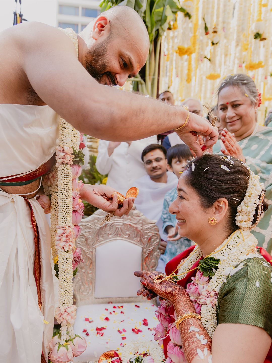 Smiling groom places flowers in brides hair during colorful South Indian Hindu wedding ceremony with guests watching.