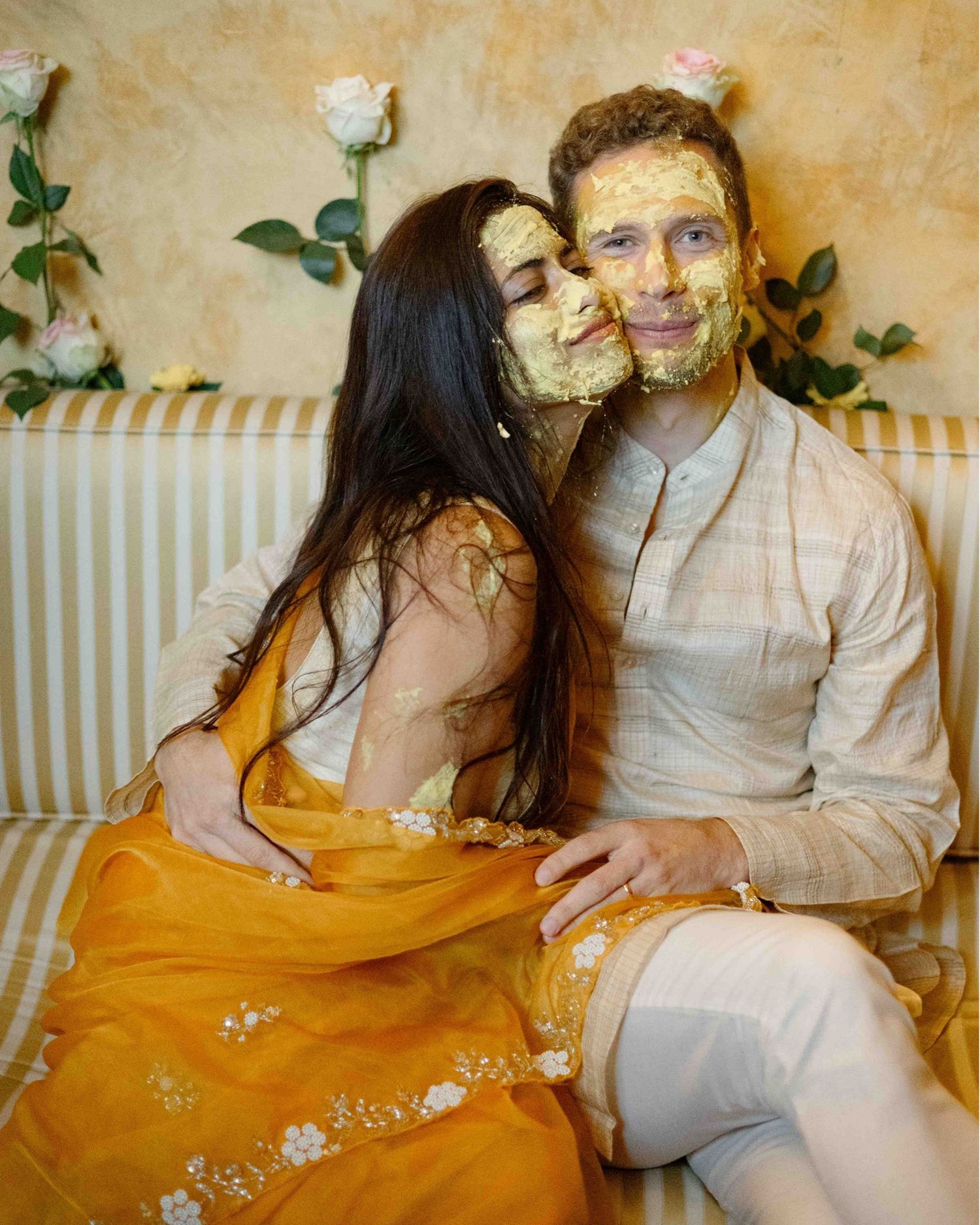 Couple covered in haldi paste during Indian wedding ceremony, bride in yellow lehenga kissing groom on cheek.