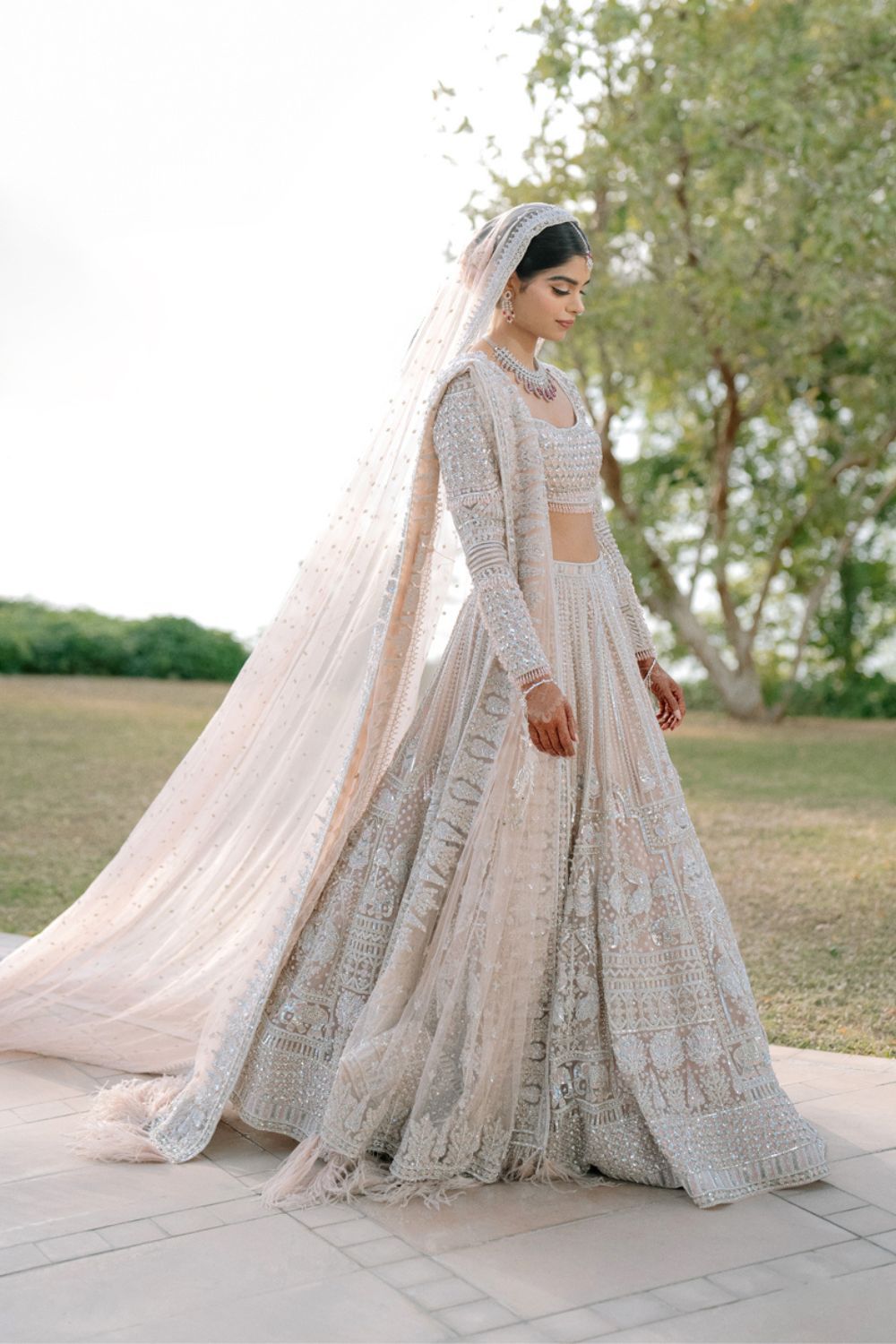 Elegant Indian bride in white embroidered lehenga with flowing veil walks gracefully outdoors.