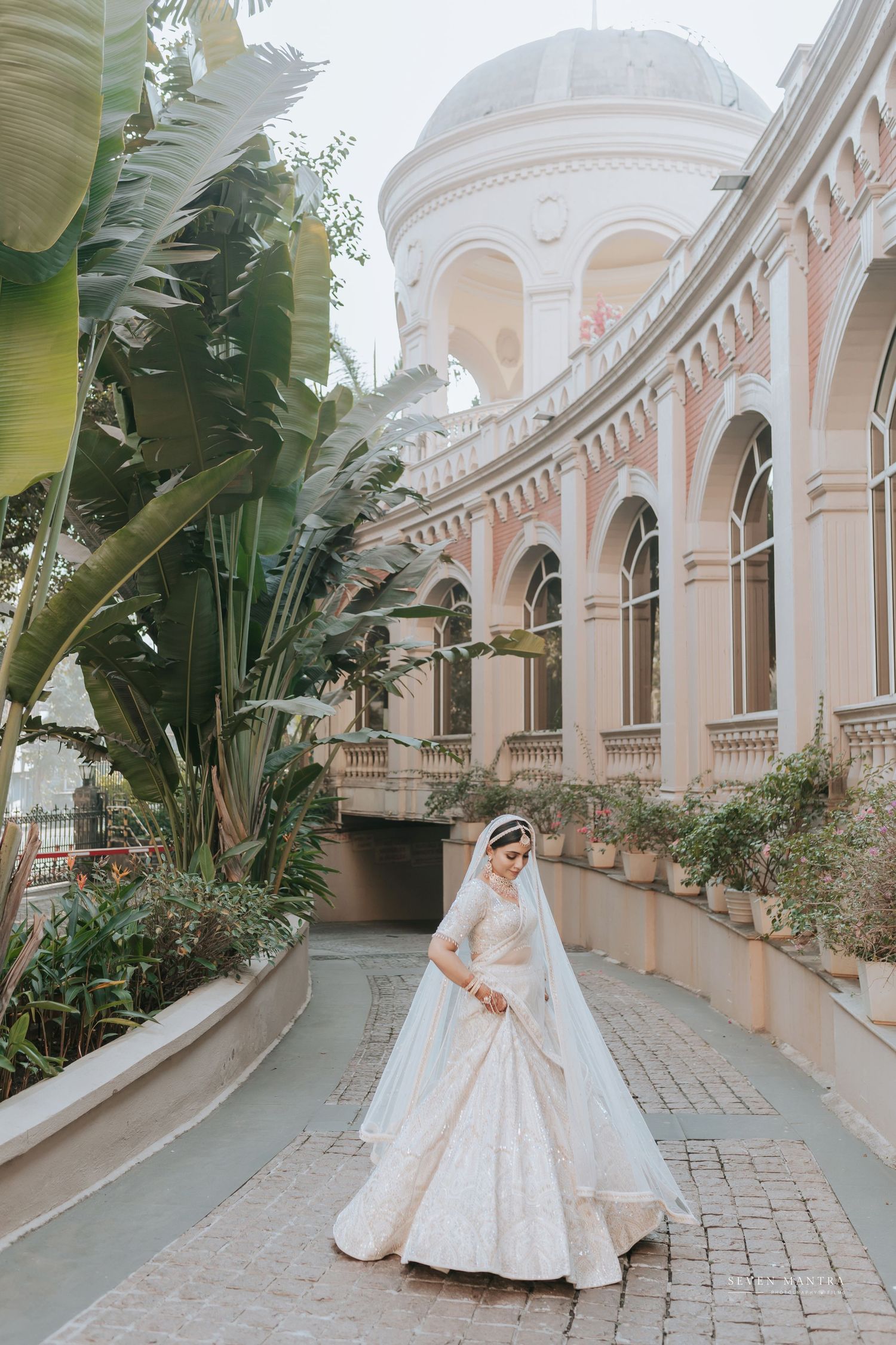 Bride in white lehenga with veil poses before a grand pink colonial building with lush tropical greenery.
