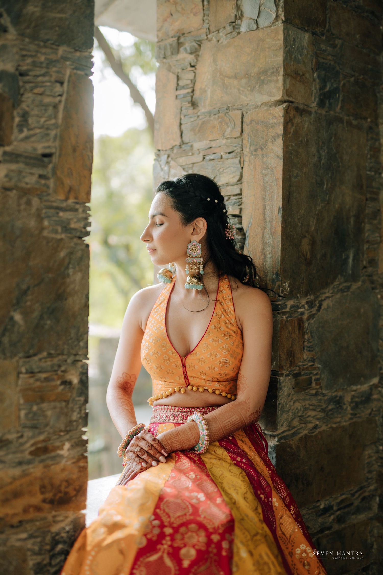 Bride in orange and red lehenga with chandelier earrings poses gracefully against rustic stone wall backdrop.