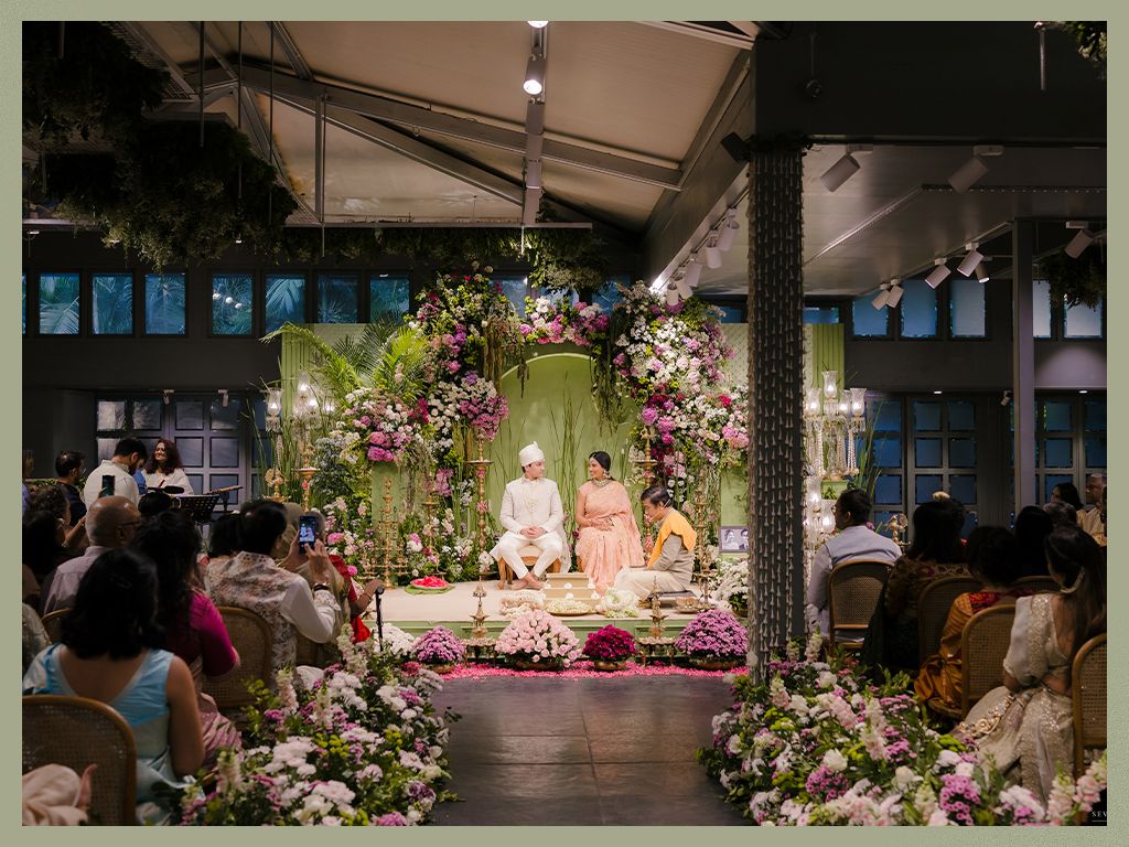 Elegant Indian wedding ceremony with lush floral arch, guests seated in decorated indoor venue.