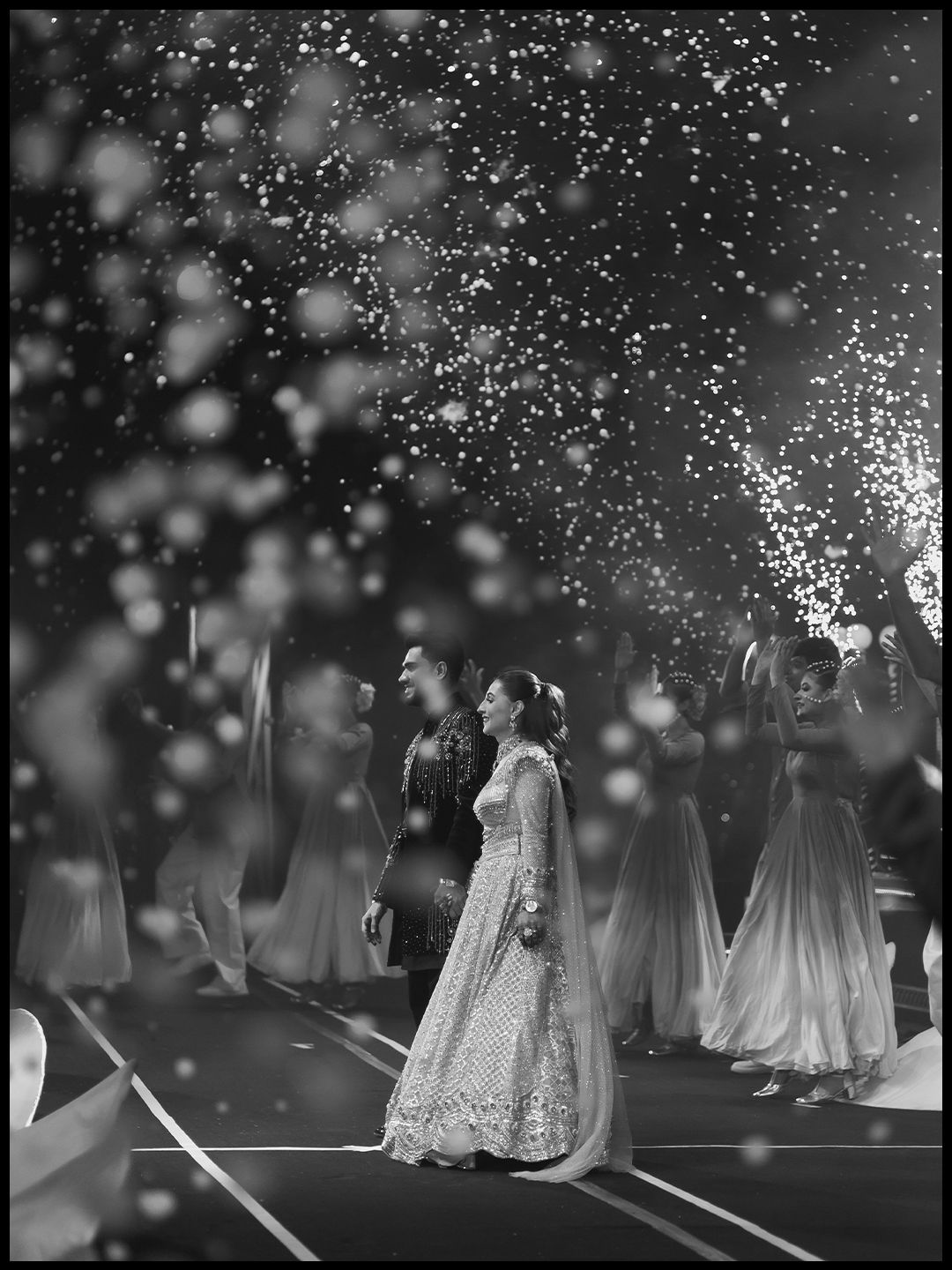 Dramatic black and white wedding entrance photo with couple surrounded by sparkling confetti and dancers.