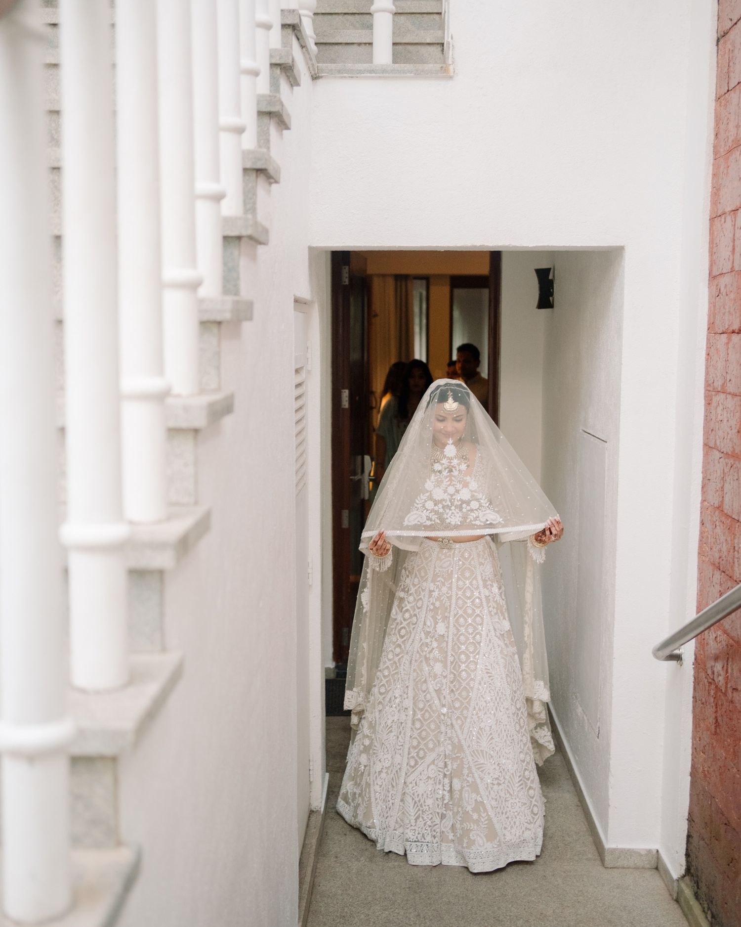Indian bride in white embroidered lehenga and veil stands in architectural doorway.