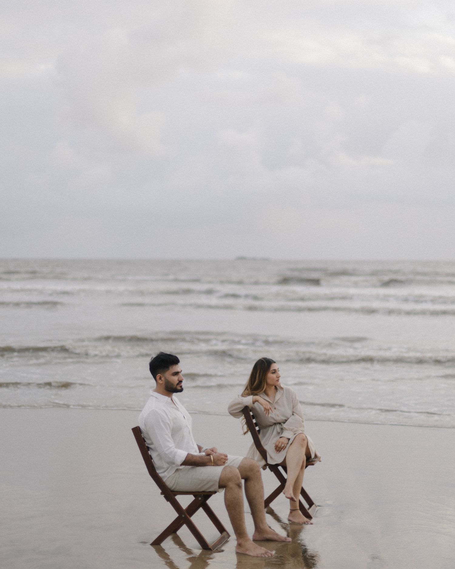 Couple sitting on wooden chairs on a cloudy beach, dressed in neutral tones with waves behind them.