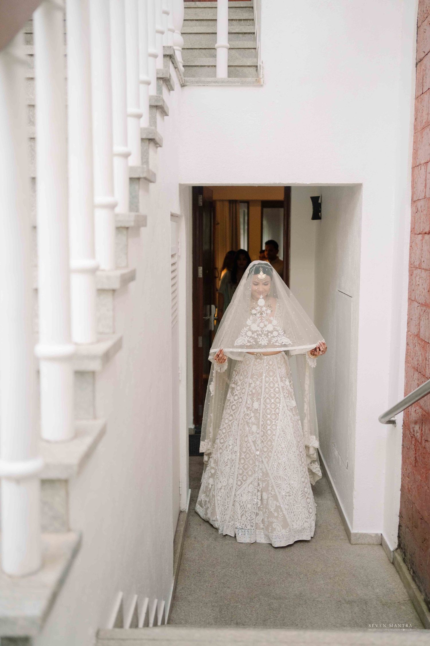 Bride in ornate white lehenga holding veil in narrow stairwell before wedding ceremony.