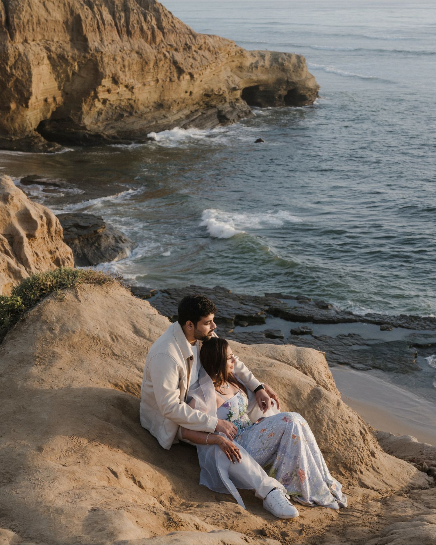 Couple sitting together on coastal cliffs overlooking scenic ocean waves at golden hour.