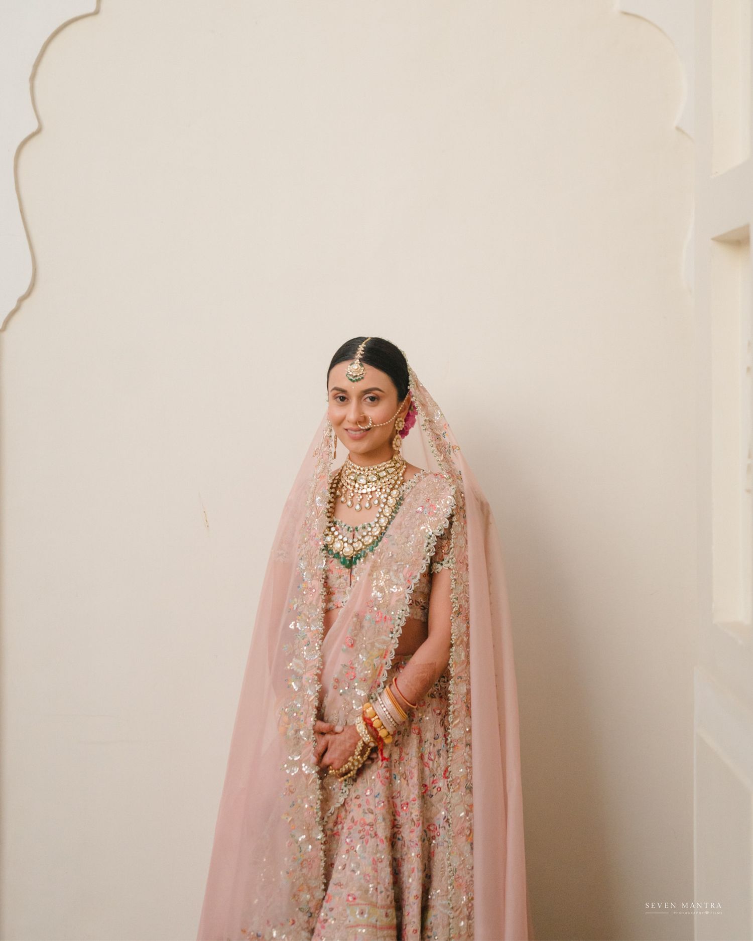Elegant Indian bride in pink lehenga with dupatta and kundan jewelry smiling softly against white wall.