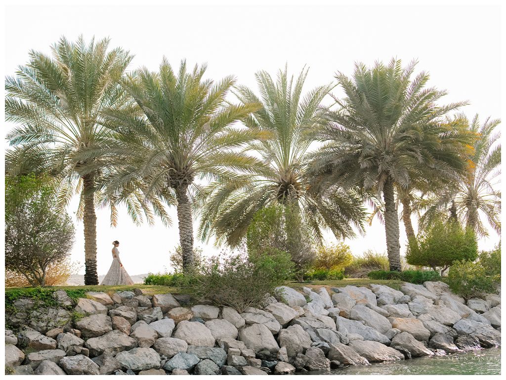 Bride in white lehenga stands among tall palm trees on rocky shoreline at outdoor Indian wedding.