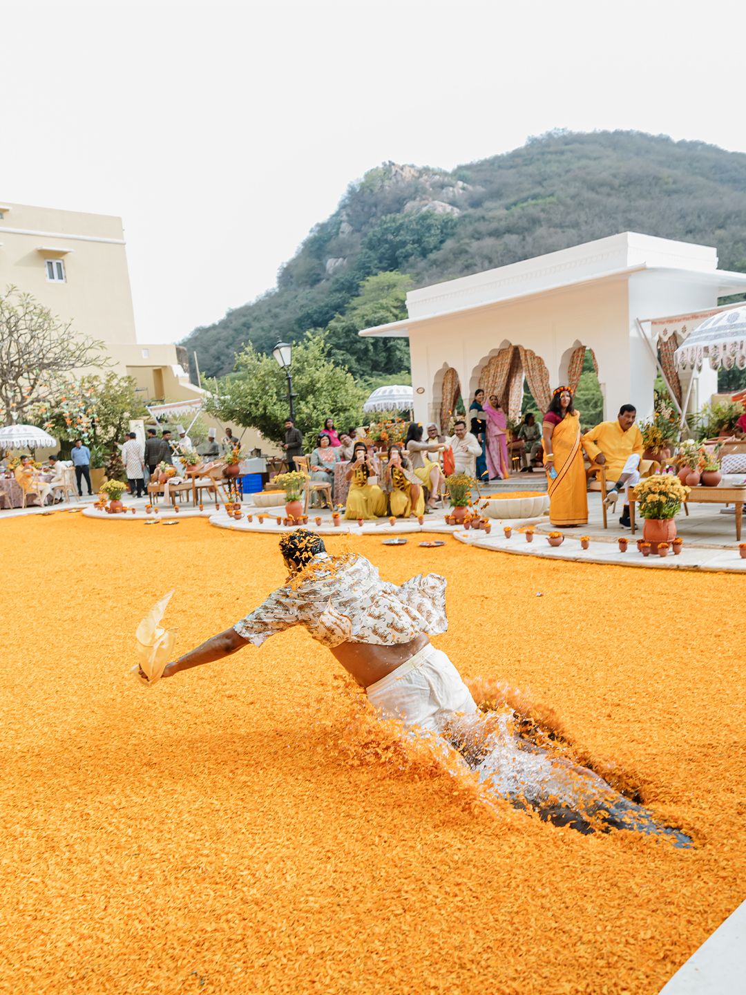 Joyful wedding guest dramatically slides through orange marigold petals at colorful outdoor Indian wedding celebration.