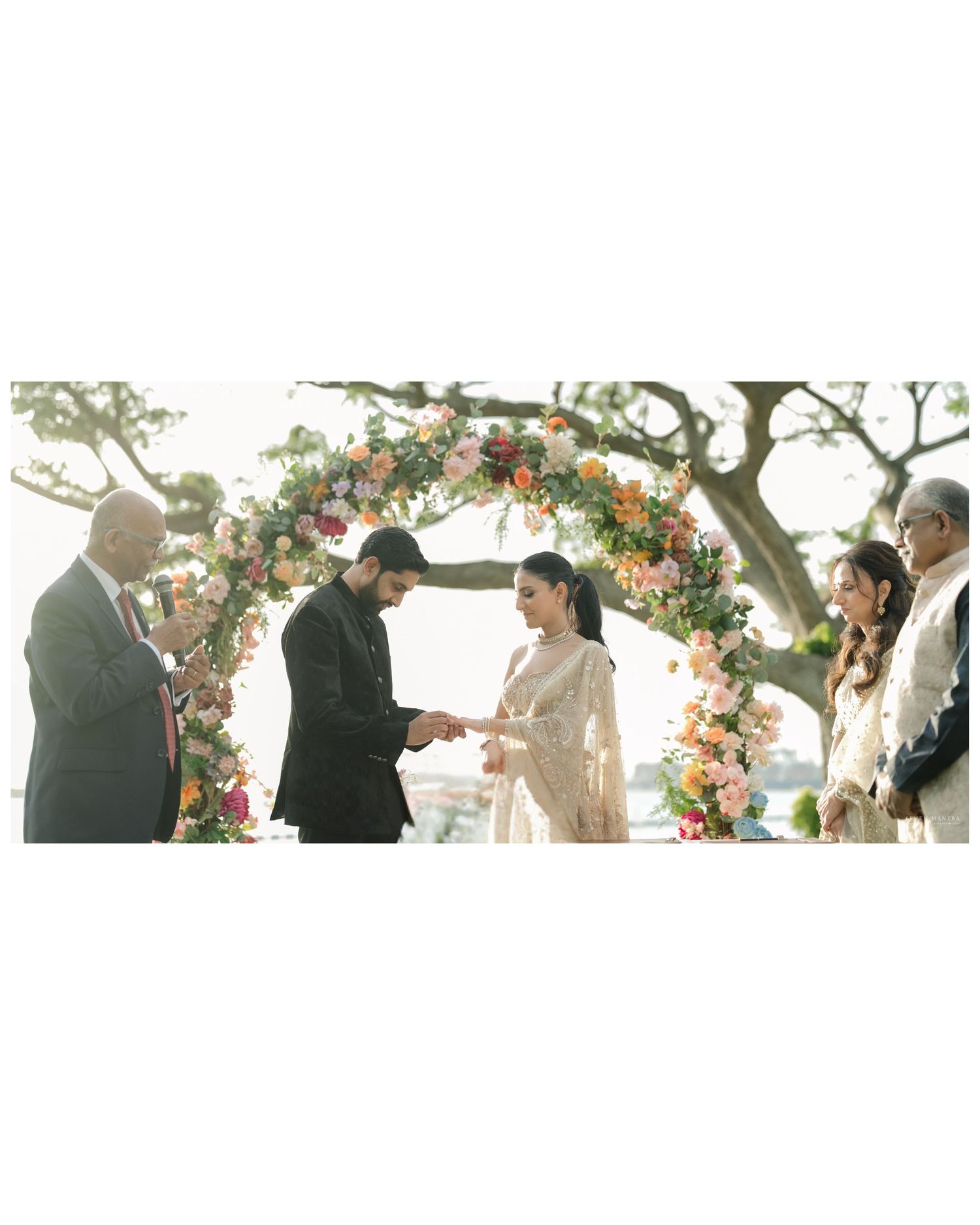 Couple exchanges rings under a colorful floral arch at an outdoor Indian wedding ceremony.