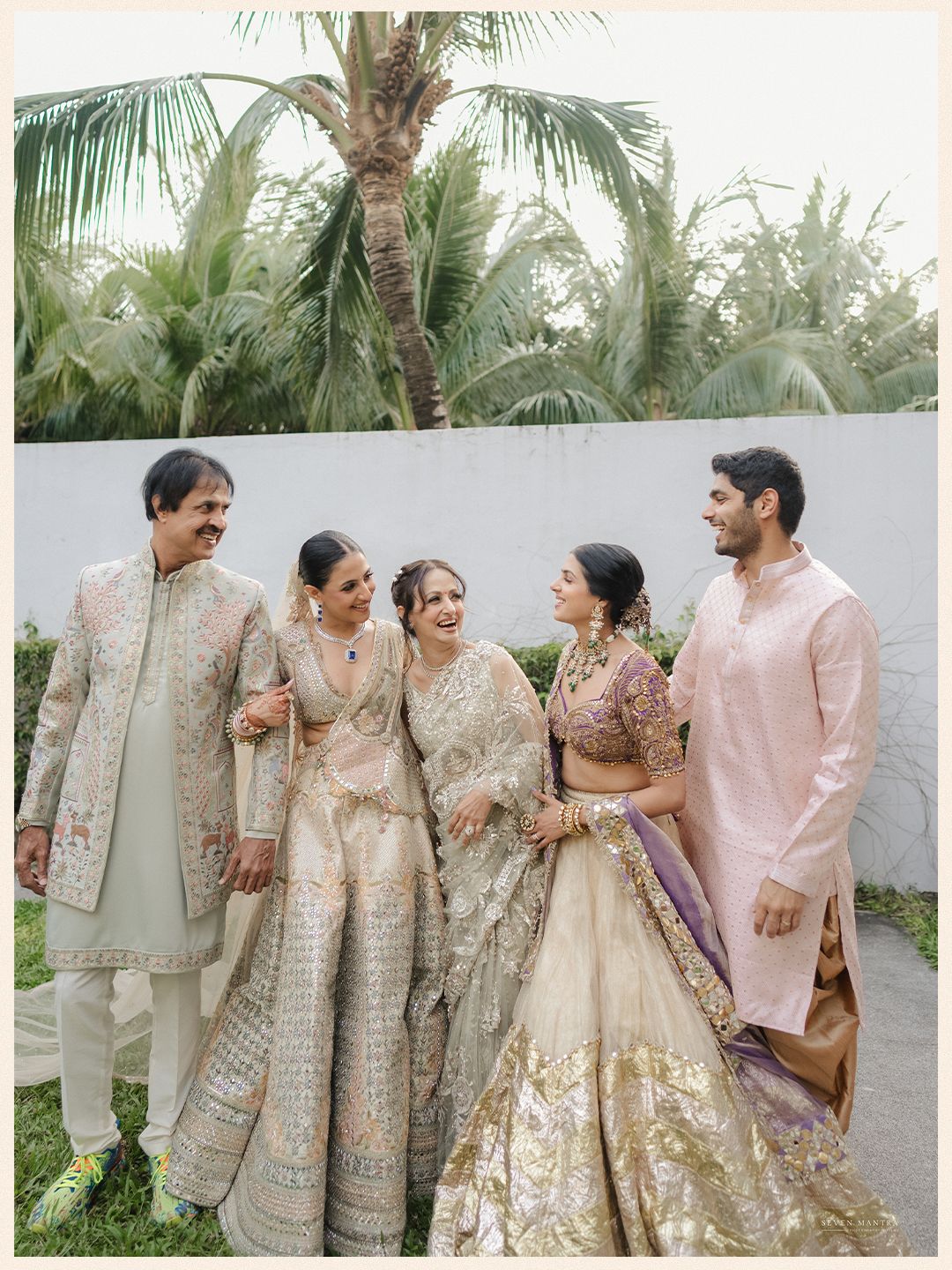 Indian wedding family portrait with bride in gold lehenga and groom in pink kurta among palm trees.