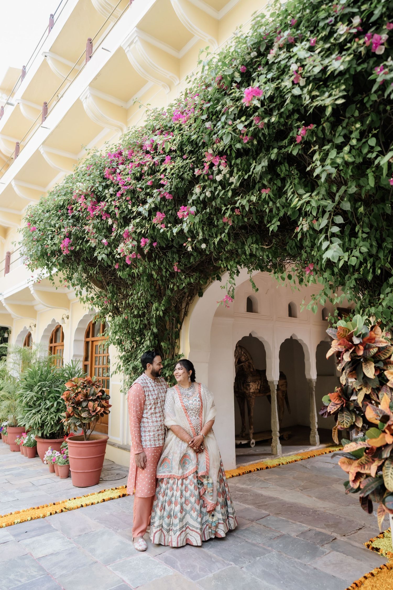 Indian couple in traditional attire posing under blooming bougainvillea at ornate palace courtyard venue.