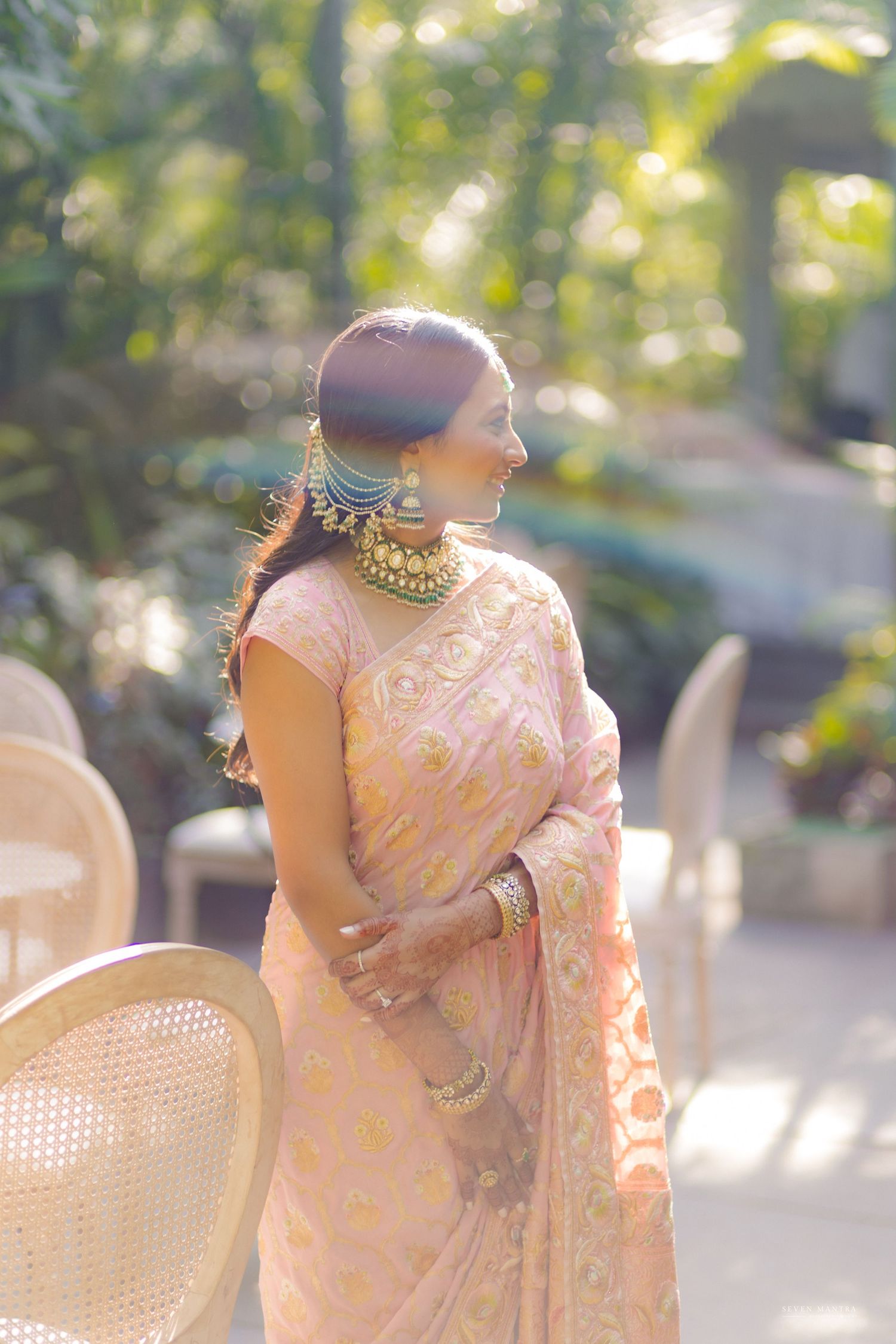 Indian bride in pink saree with statement kundan jewelry posing outdoors in golden sunlight.