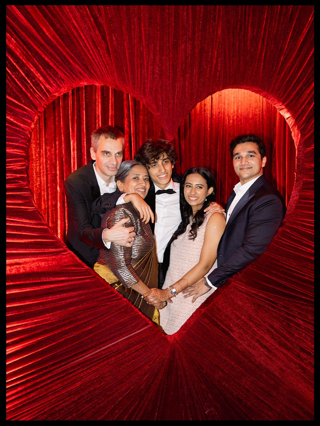 Happy family of five posing inside heart-shaped red fringe photo booth at wedding reception.