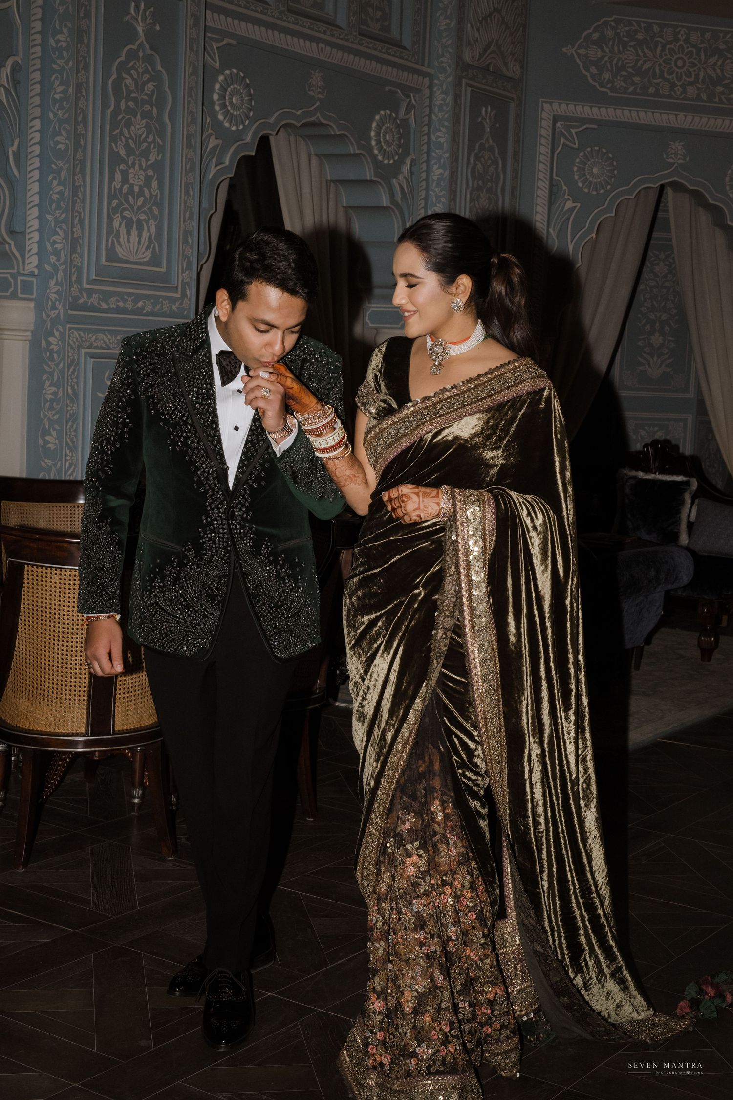 Couple in elegant Indian wedding reception outfits, groom kissing bride's hand in ornate venue.