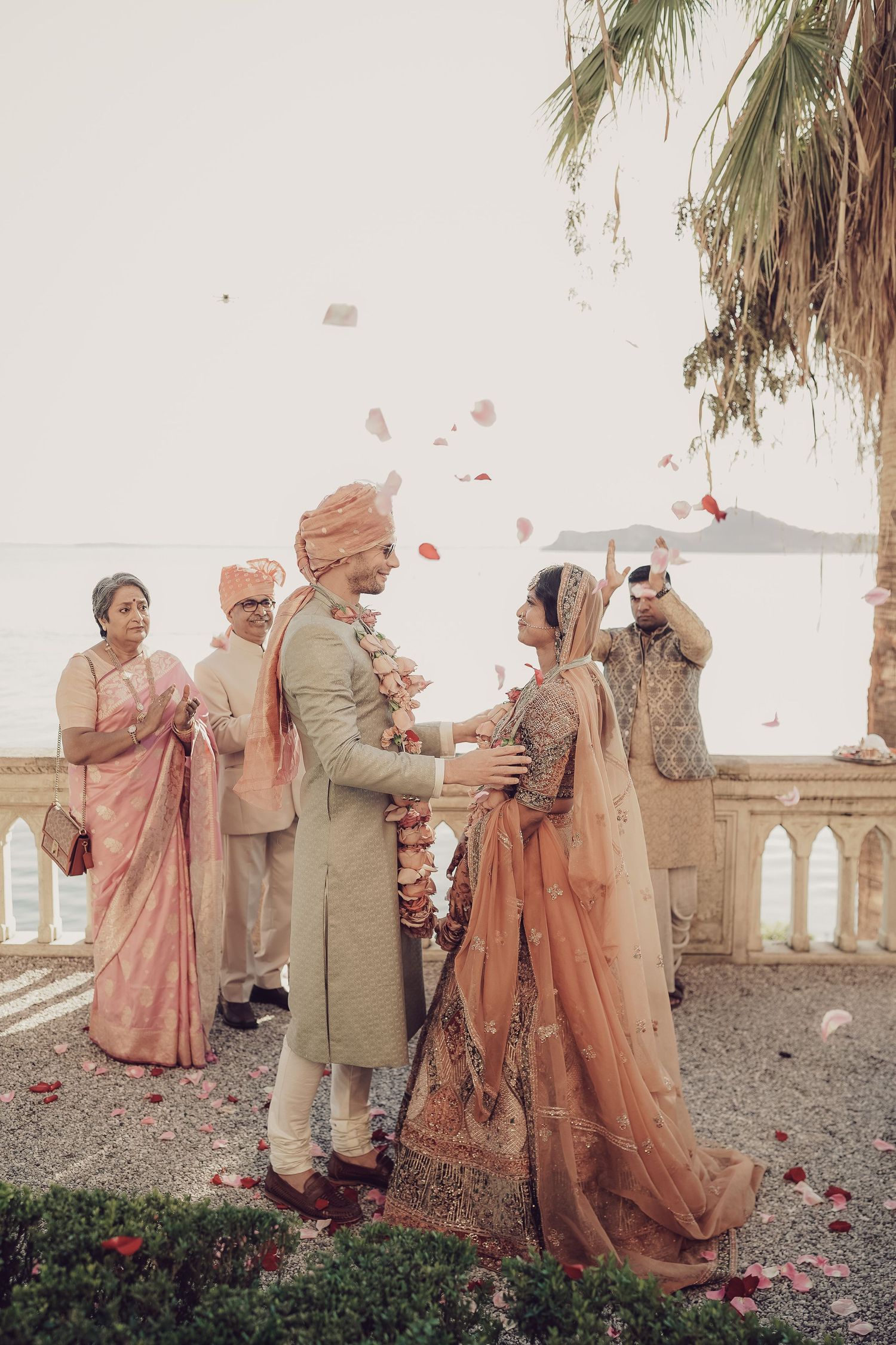 Indian bride and groom exchange garlands as rose petals shower them at lakeside wedding ceremony.