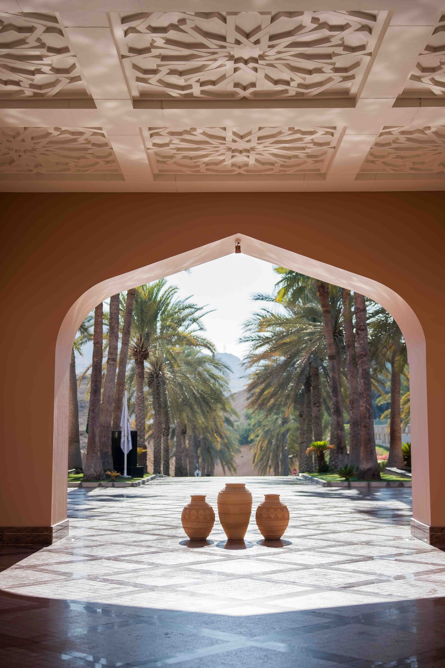 Elegant Moroccan archway with ornate ceiling, terracotta vases, and palm tree-lined pathway beyond.