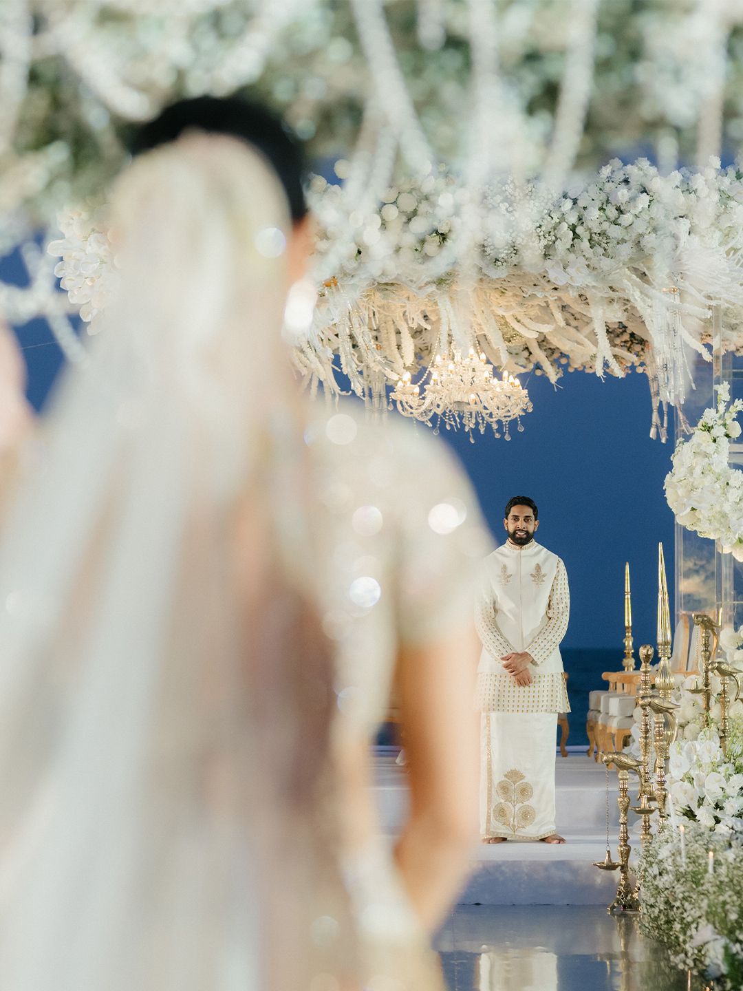 Bride in gold lehenga walks toward groom waiting under stunning white floral mandap at beach wedding.