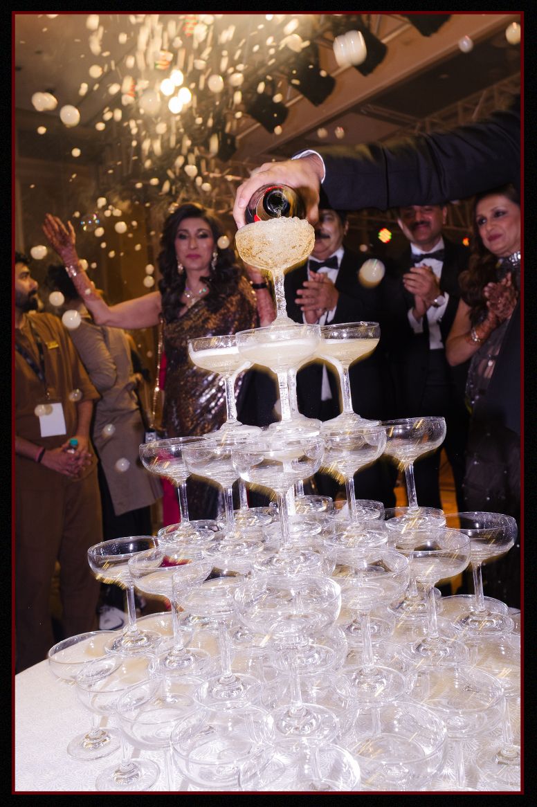 Champagne tower being filled at elegant Indian wedding reception with guests celebrating in background.