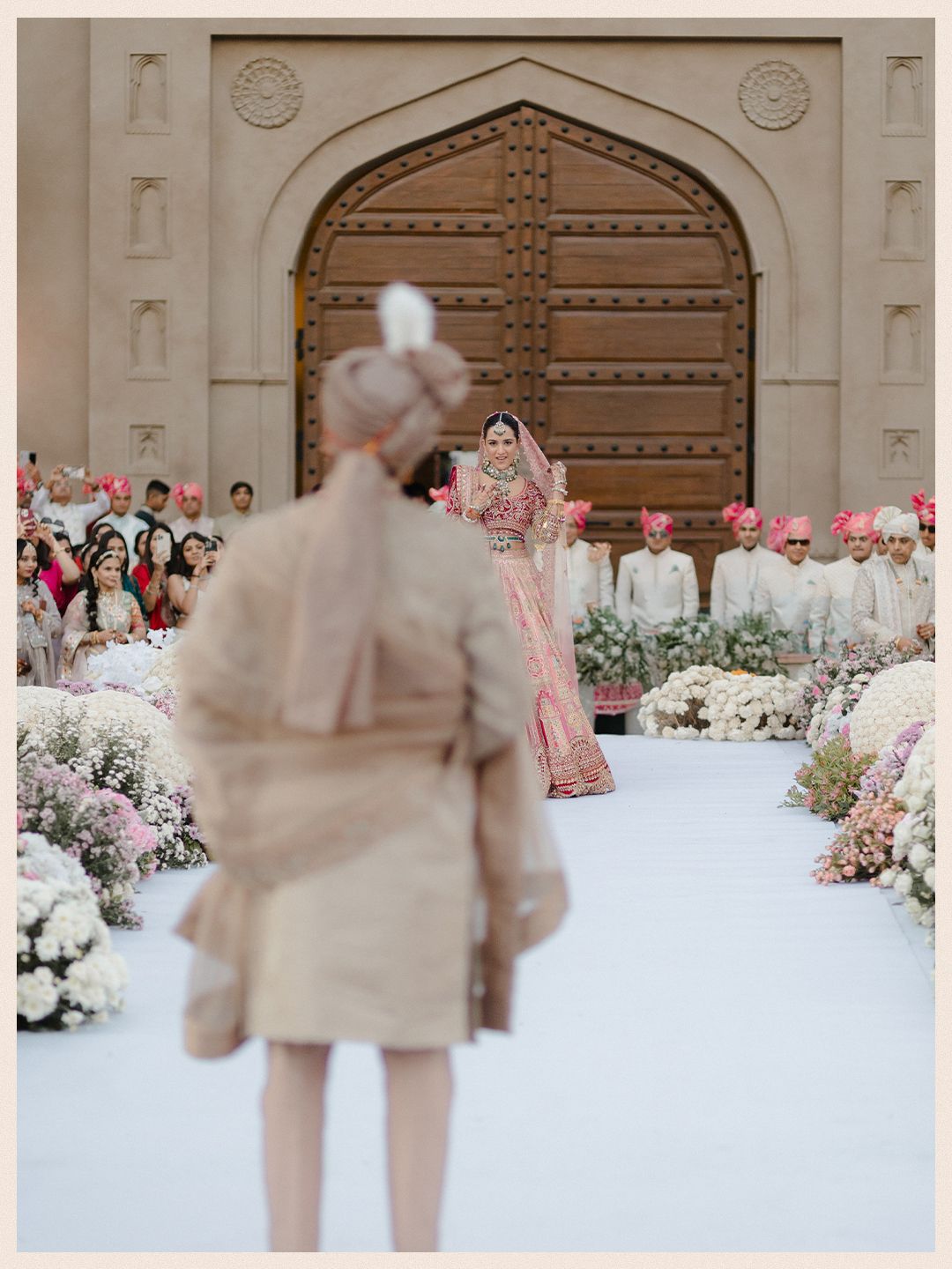 Indian bride in pink lehenga walking down white floral aisle toward groom at grand arched wedding venue.