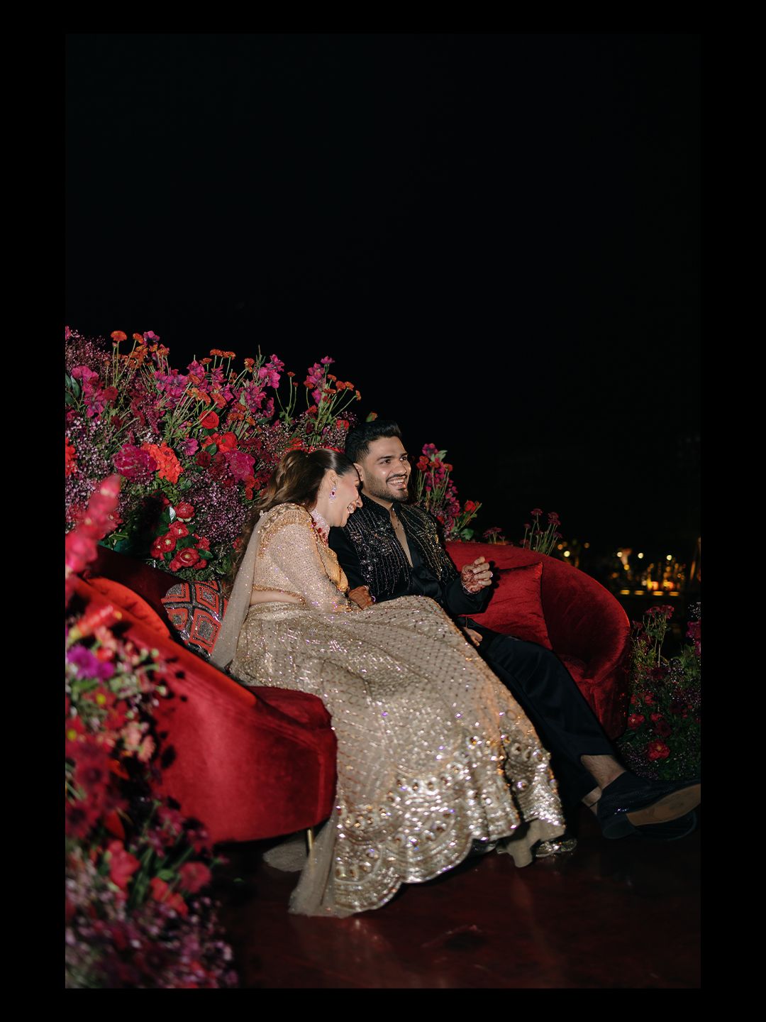 Bride and groom sitting together on red velvet sofa surrounded by vibrant pink floral arrangements at night.