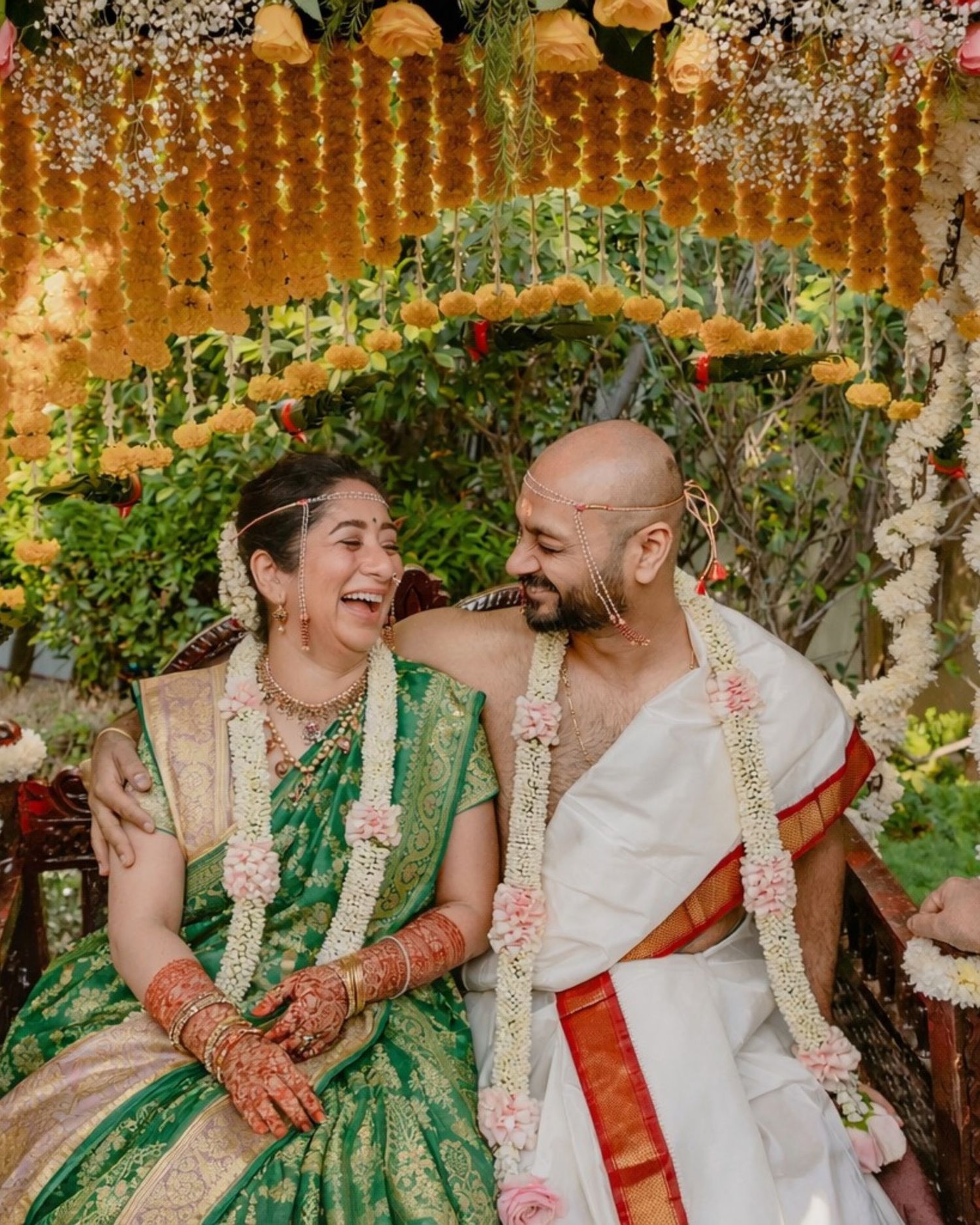Joyful Indian bride in green silk saree and groom in white dhoti laughing together under floral wedding mandap.