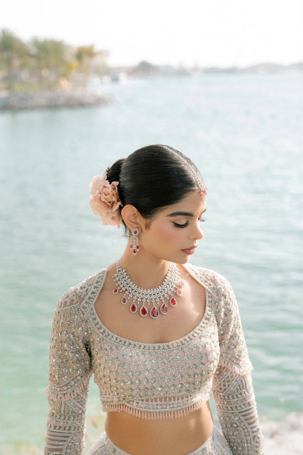 Indian bride in embroidered blouse with ruby and diamond necklace poses near turquoise water.