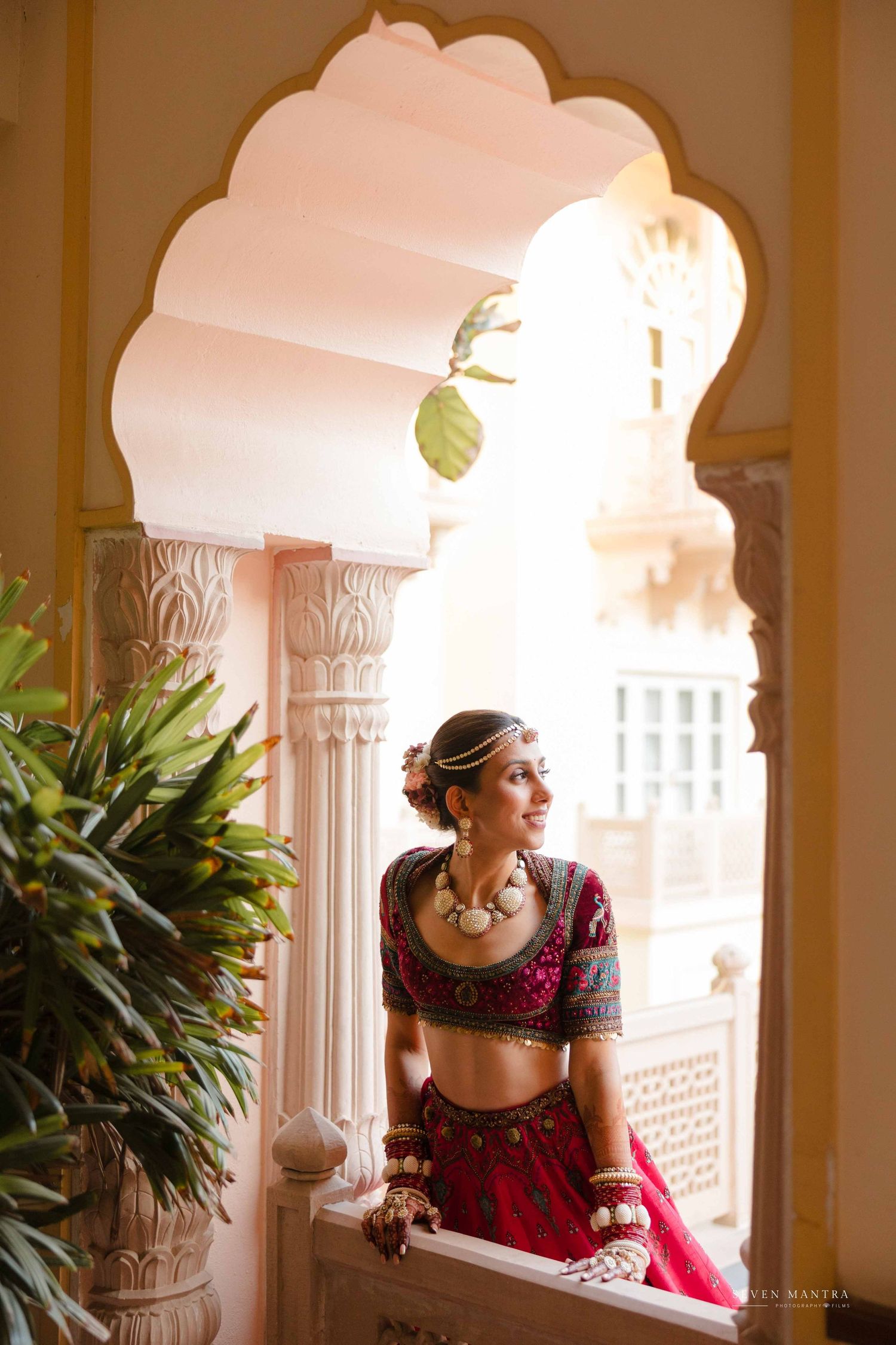 Bride in crimson lehenga with silver jewelry posing gracefully in ornate white arched palace balcony.
