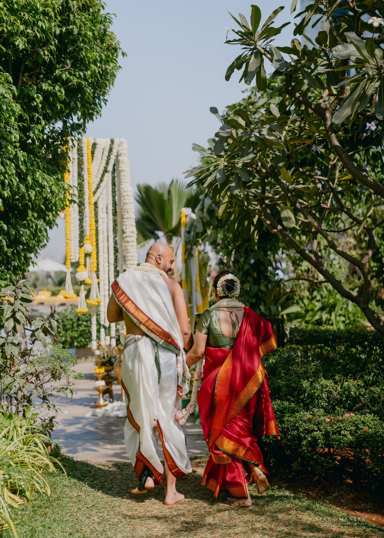 Couple in traditional South Indian wedding attire walk hand-in-hand through a lush garden decorated with marigold garlands.