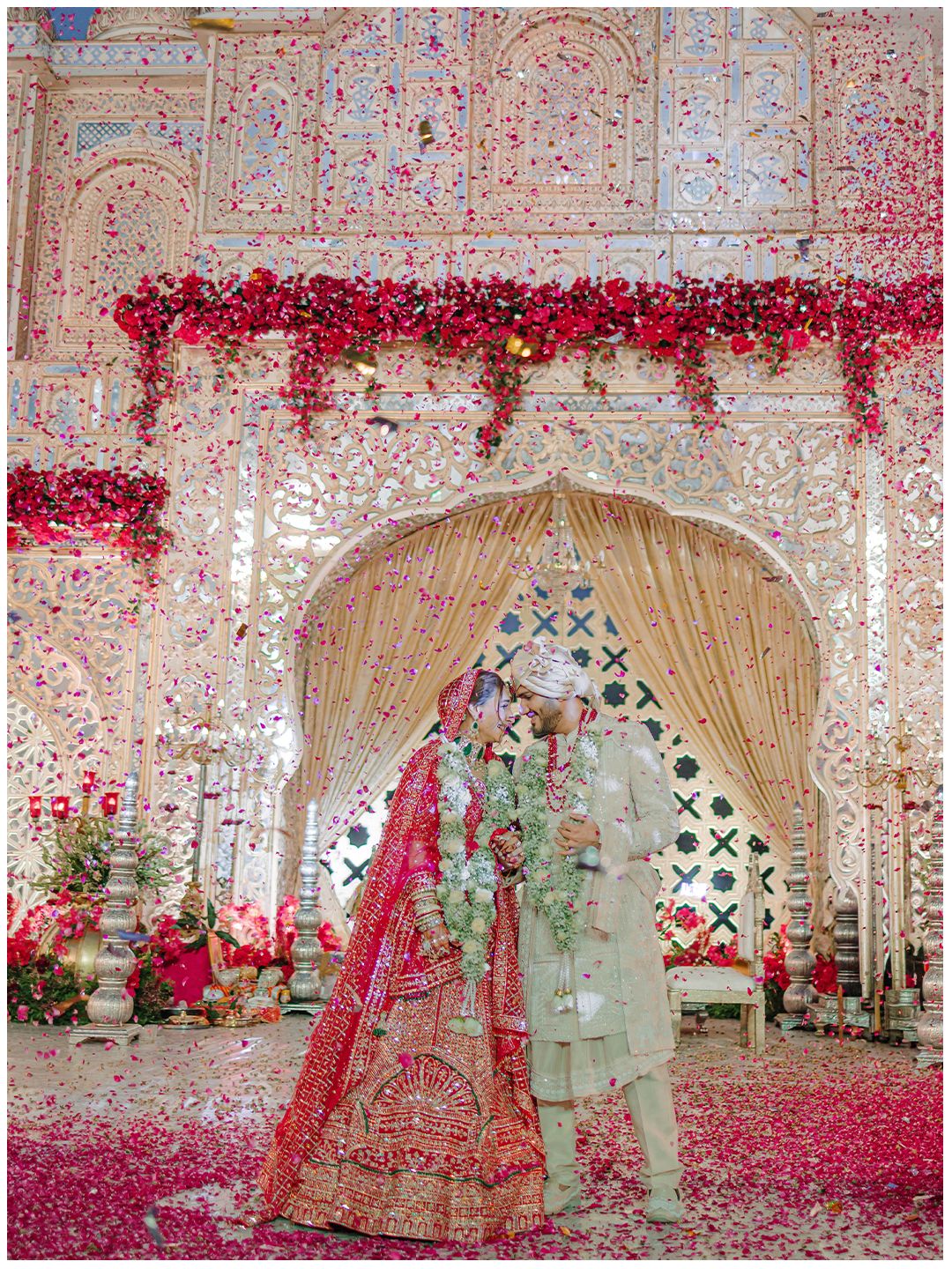 Indian bride and groom under floral arch with red rose petals falling at ornate wedding venue.