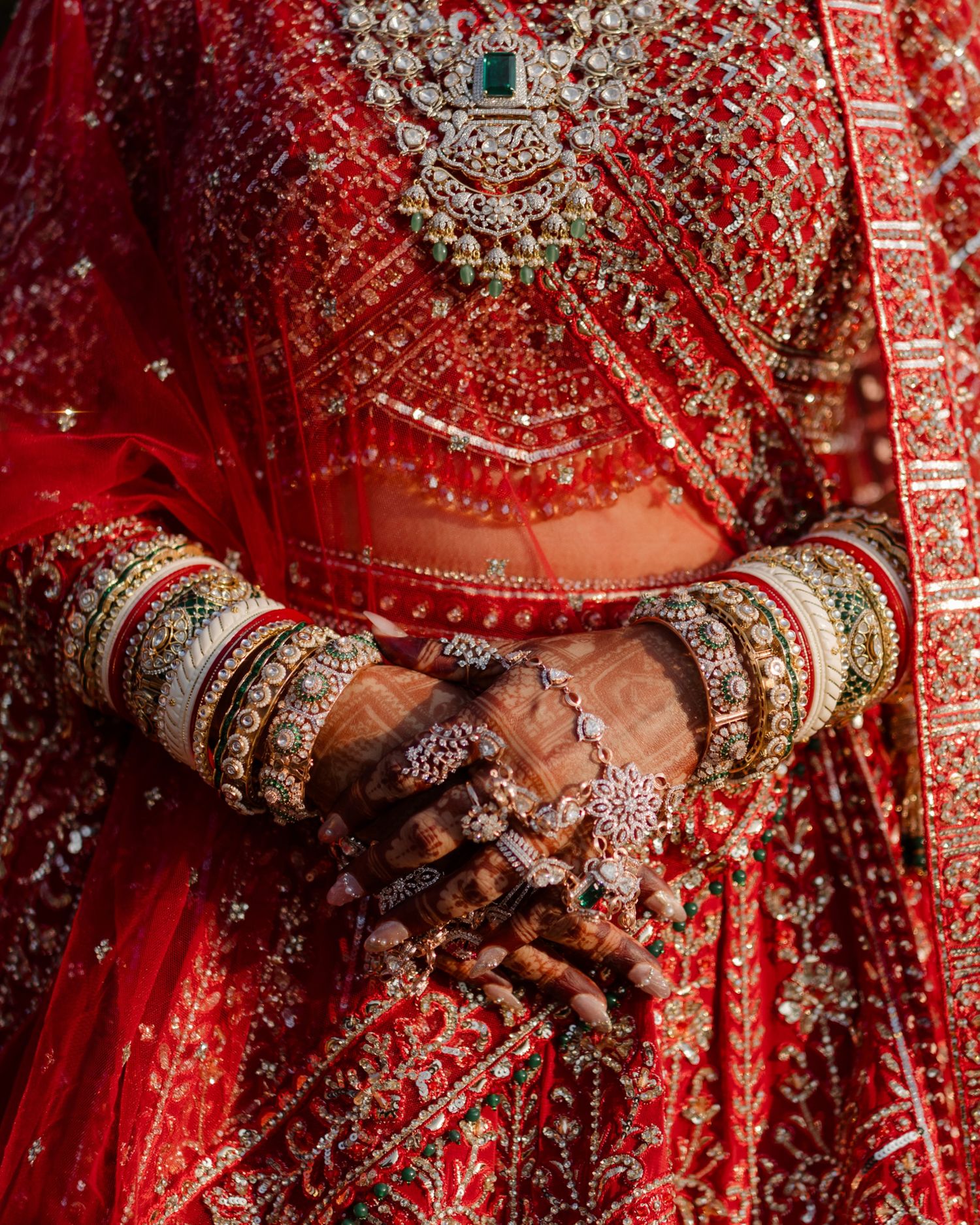 Close-up of Indian bride hands with mehndi, gold bangles, and intricate red bridal lehenga embroidery.