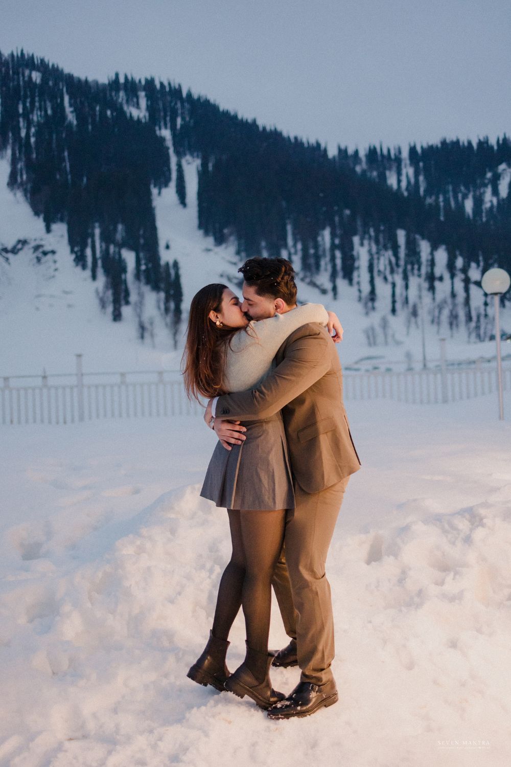 Couple shares a romantic kiss in snowy mountain landscape at golden hour.