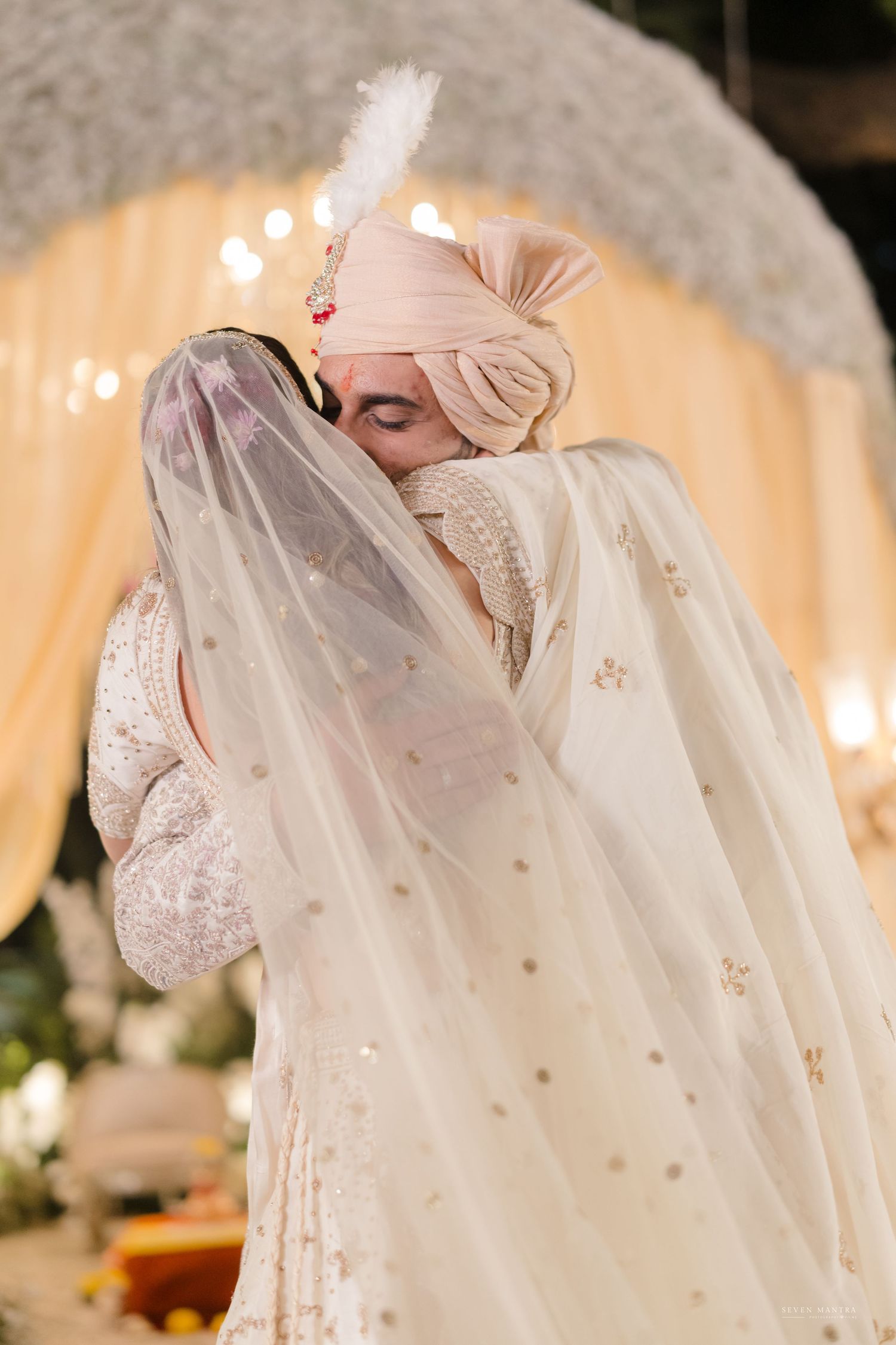 Indian groom in blush turban embraces veiled bride in embroidered lehenga before a glowing floral wedding backdrop.