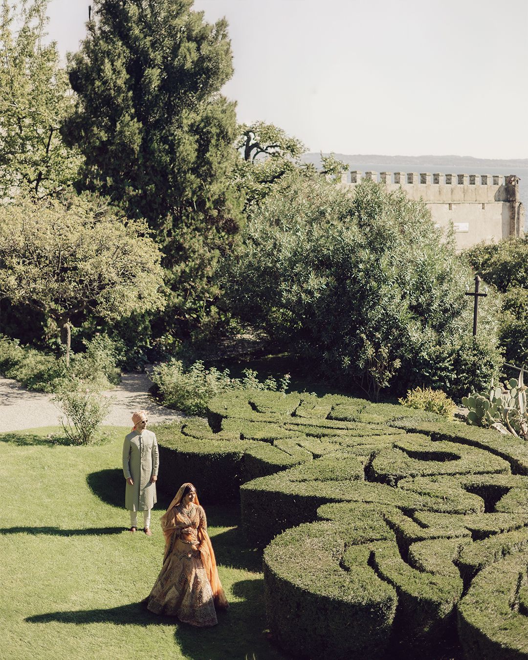 Bride and groom in traditional Indian wedding attire stroll through ornate topiary garden near historic castle.