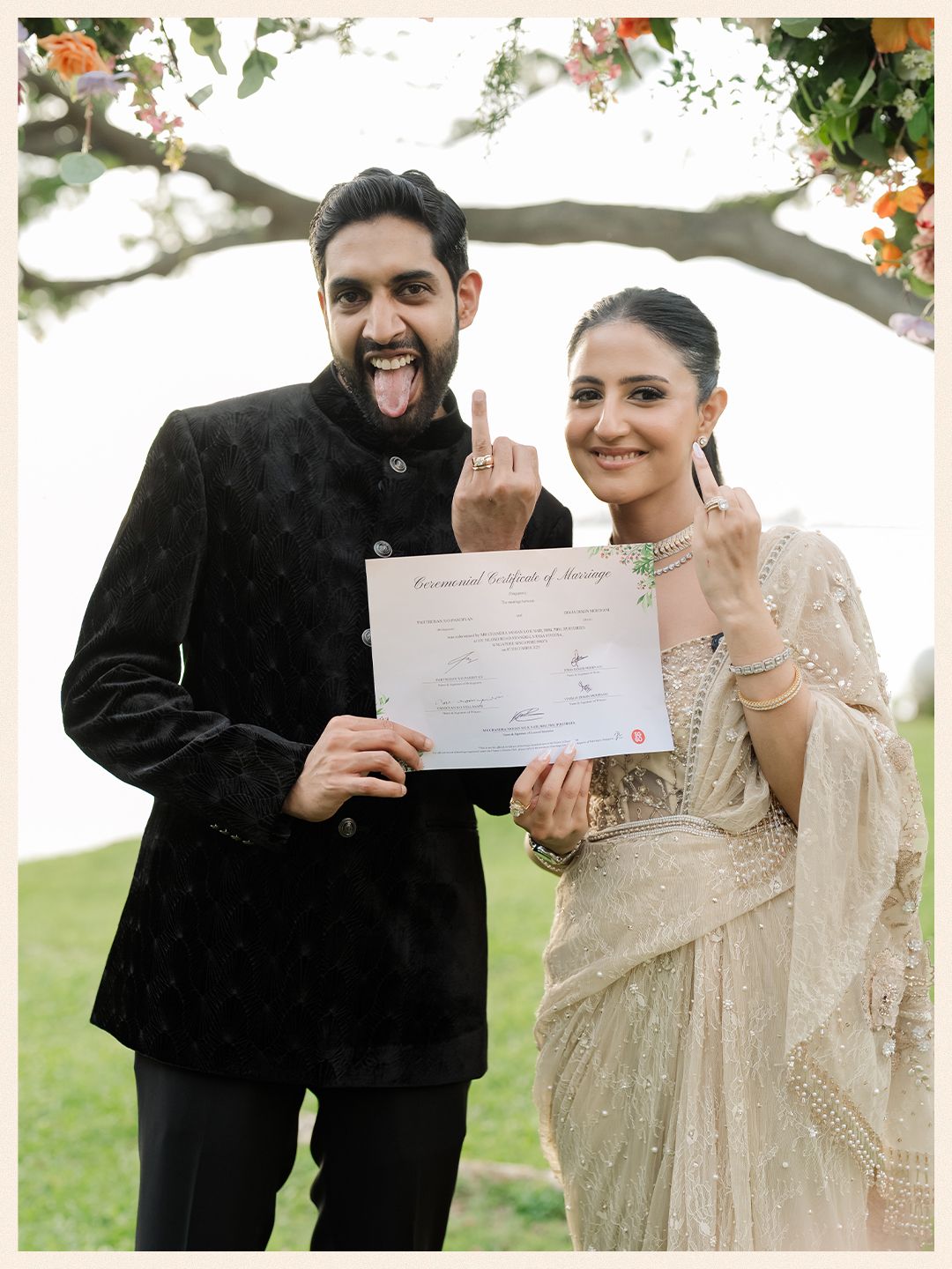 Newlyweds holding marriage certificate and showing rings while smiling under floral wedding arch.