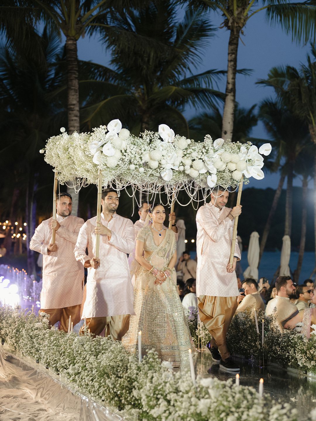 Bride enters under a floral canopy carried by groomsmen at an elegant Indian beach wedding ceremony.