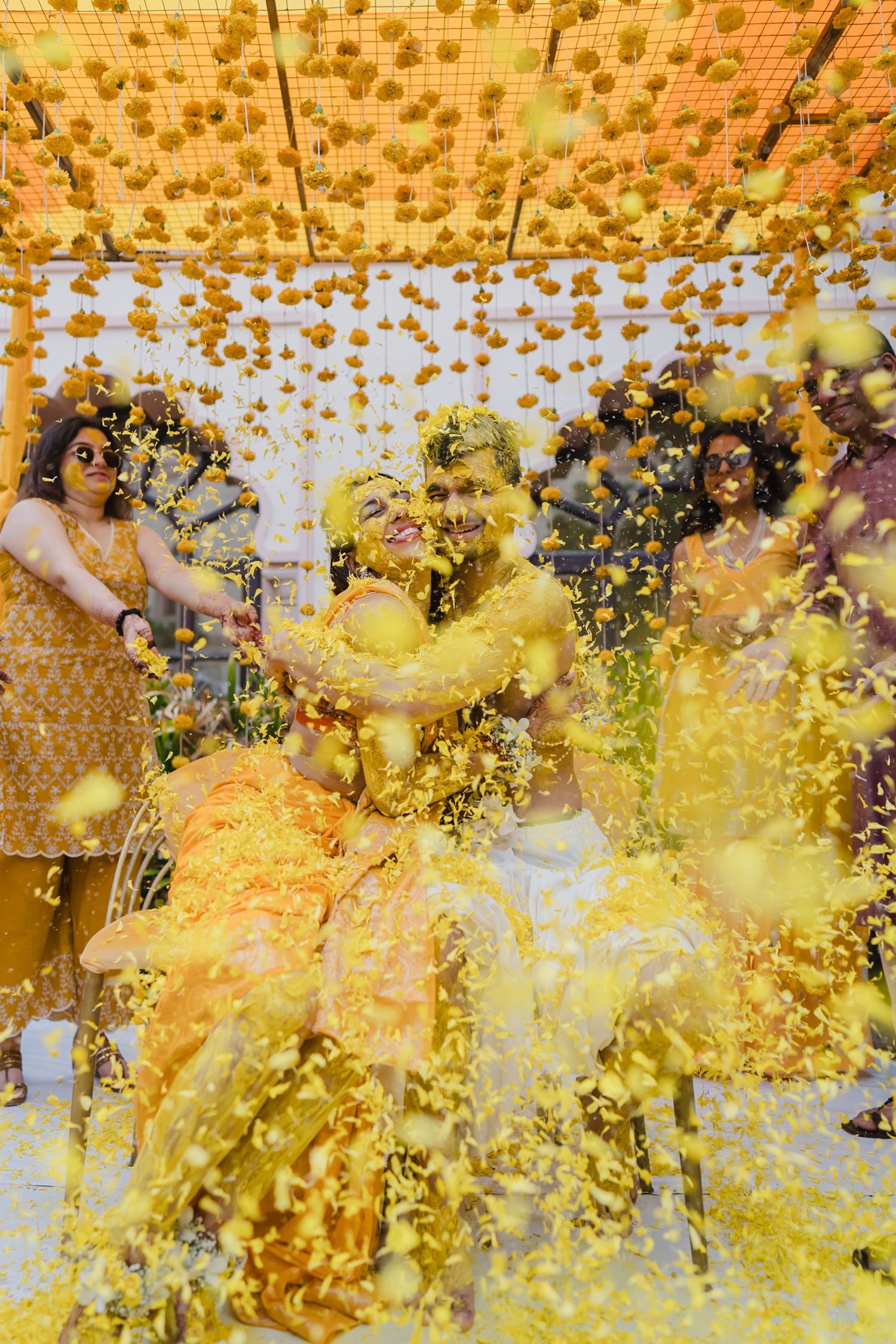 Couple covered in yellow turmeric powder during vibrant haldi ceremony with marigold flower decorations.
