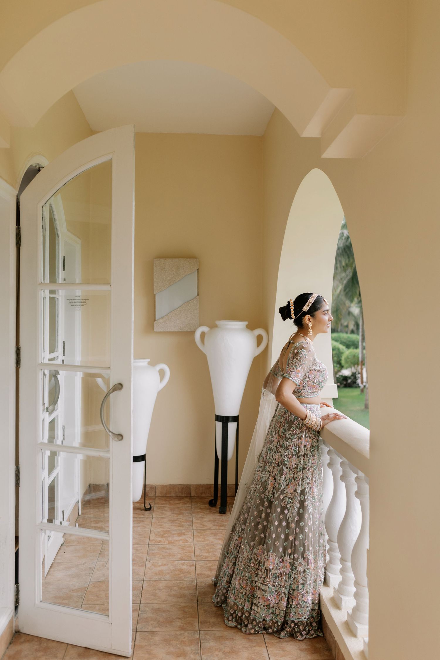 Indian bride in grey floral lehenga stands on balcony with arched doorway in soft yellow interior.